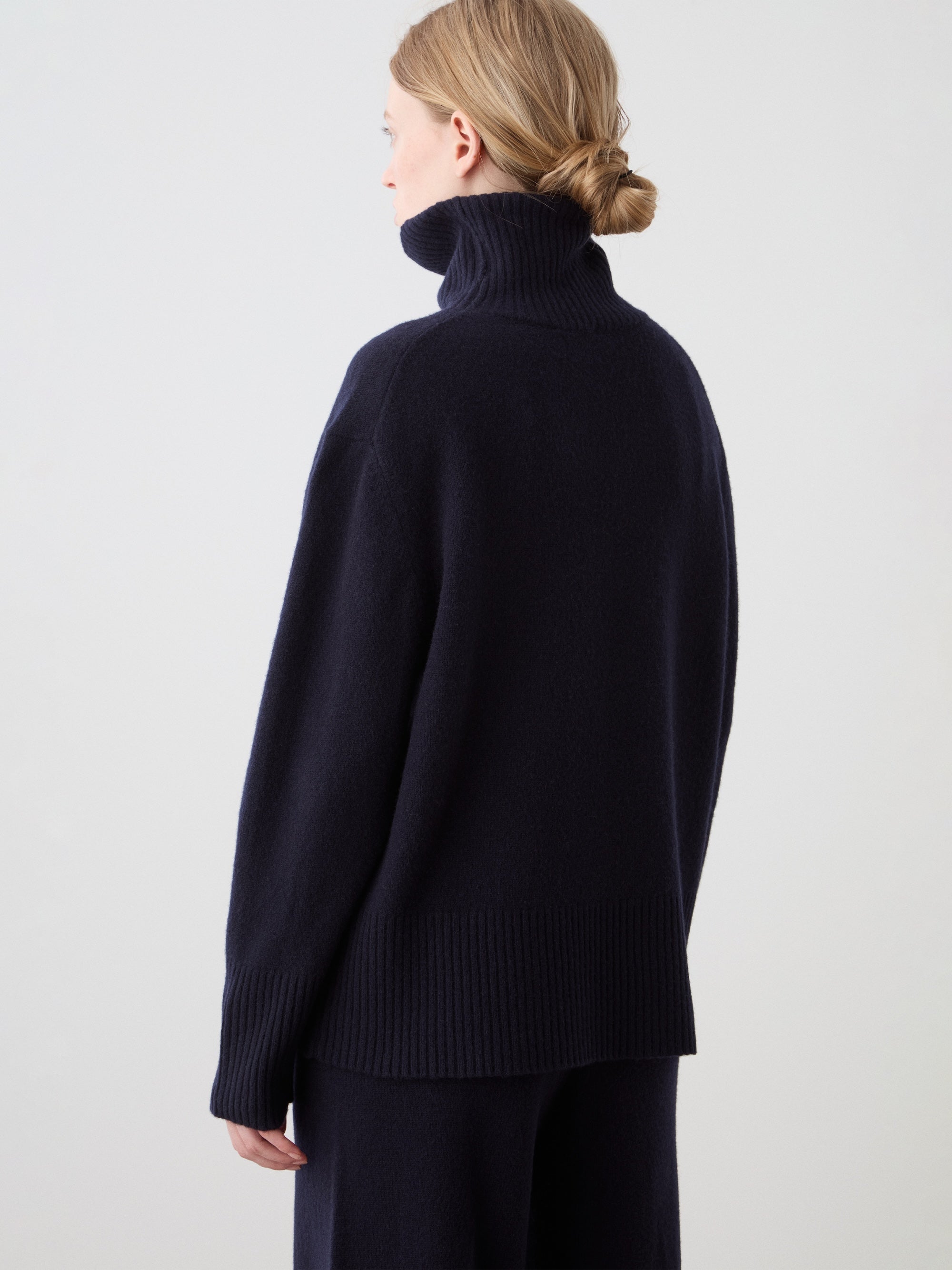 A person with blond hair in a low bun faces away from the camera against a plain light background, wearing the We Norwegians Geilo BoyFriend Sweater for women, made from Extrafine Merino Wool.