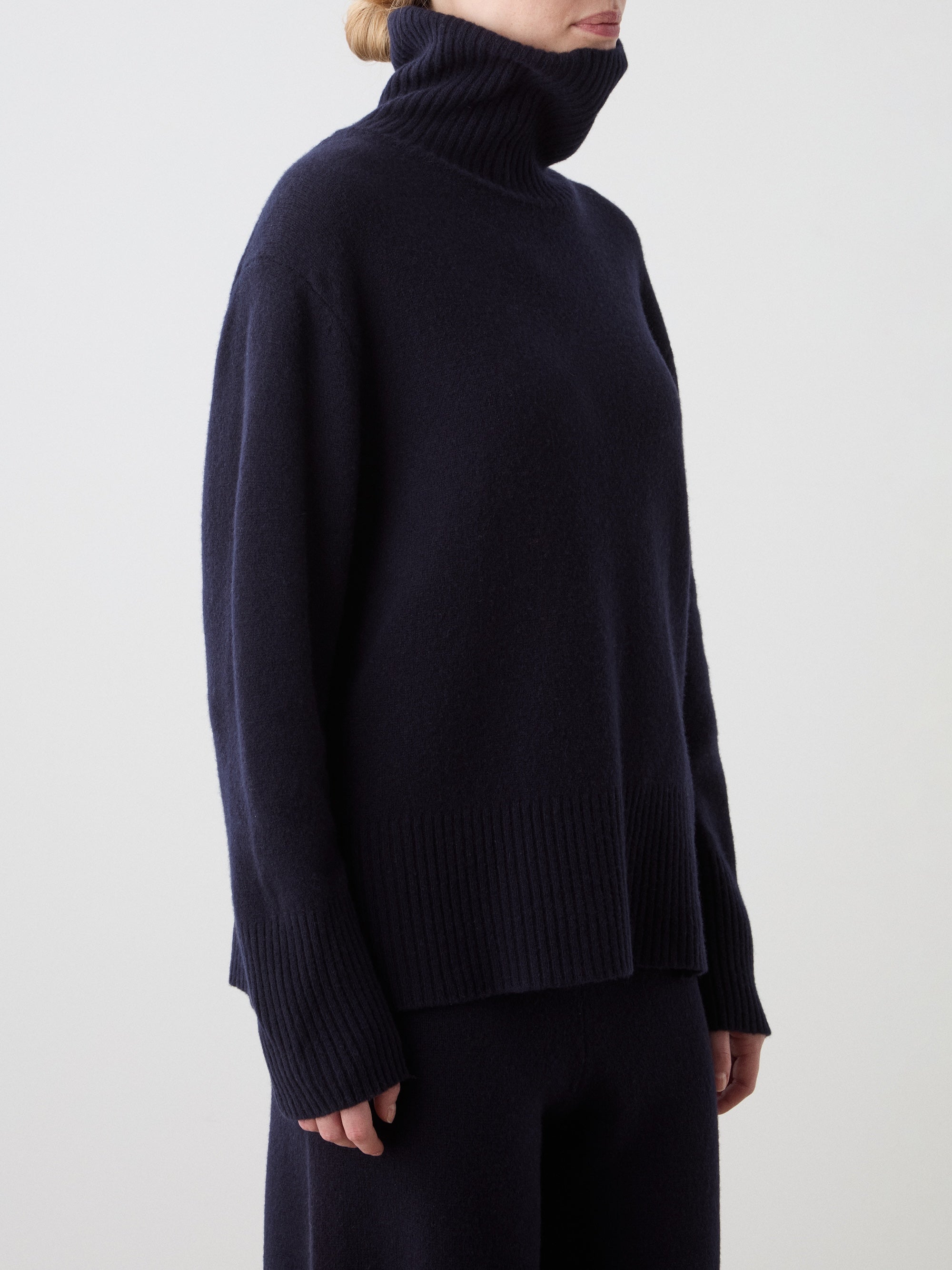 A person models the We Norwegians Geilo BoyFriend Sweater for women—a loose-fitting, dark navy turtleneck with long sleeves, ribbed cuffs and hem—against a plain light background, their face partially hidden.