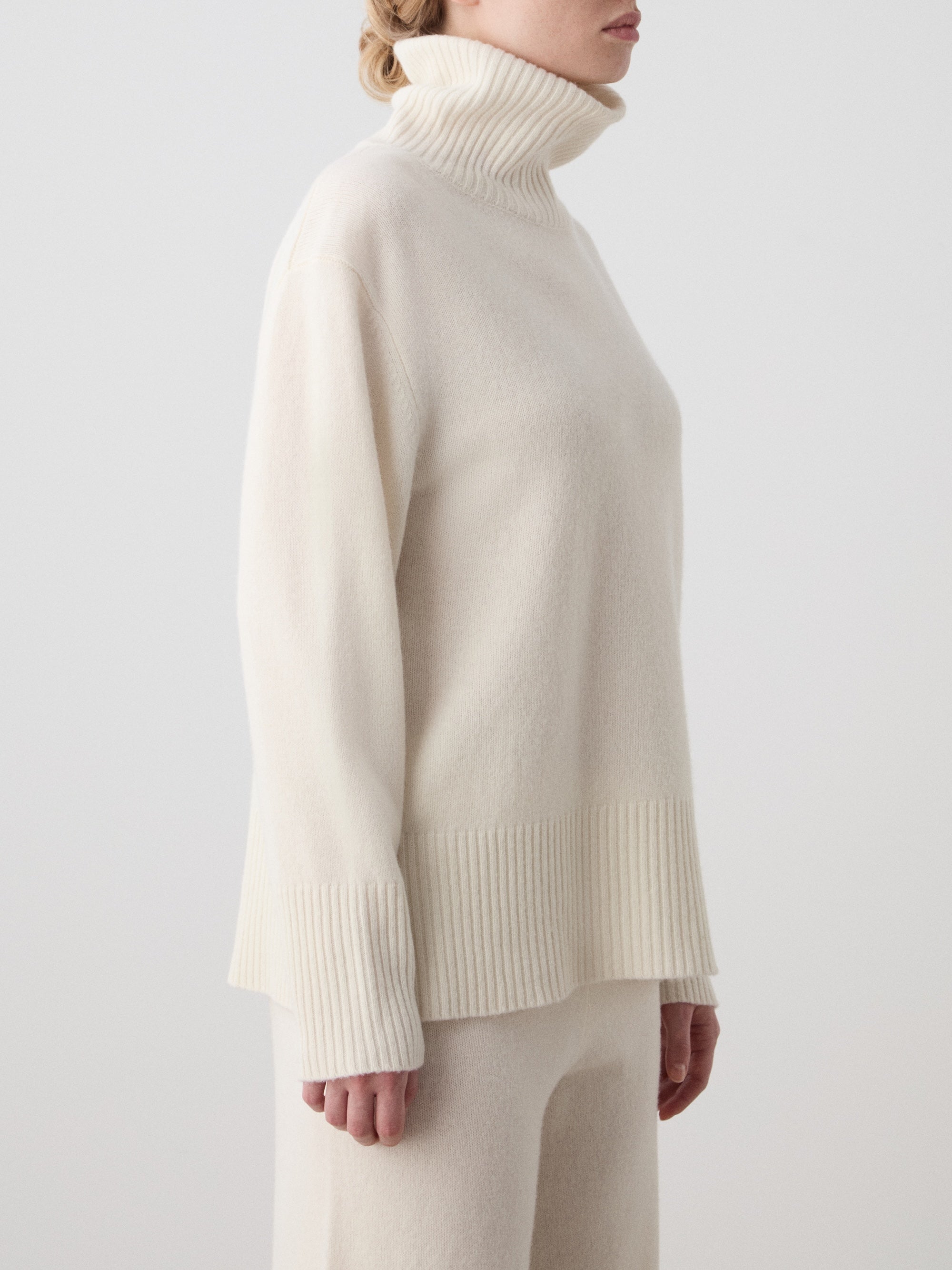 A person wears the We Norwegians Geilo BoyFriend Sweater women in cream Extrafine Merino Wool, featuring ribbed collar, cuffs, and hem, styled with matching pants against a light background.