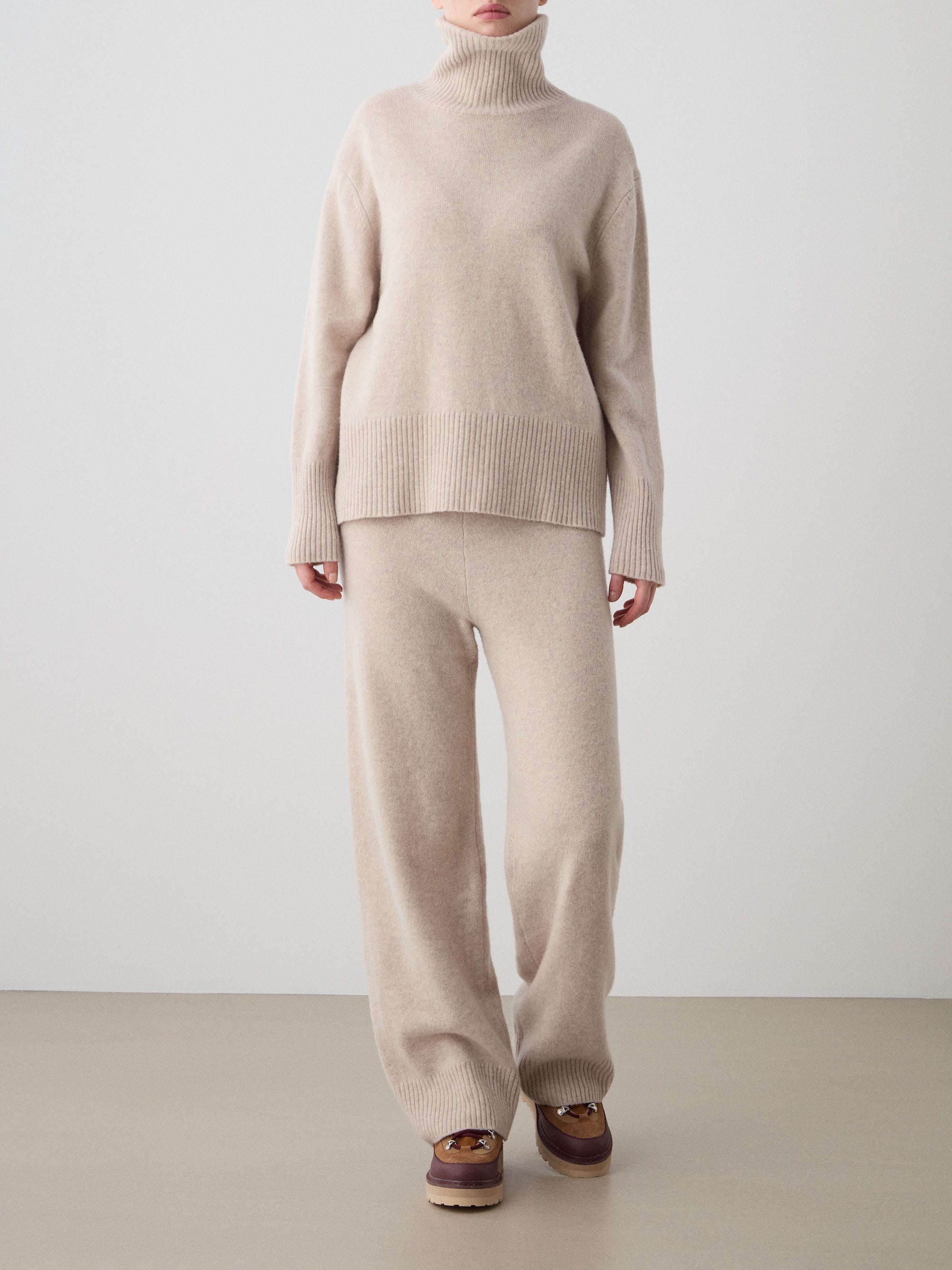 A person stands against a plain background wearing the We Norwegians Geilo BoyFriend Sweater for women, matching wide-leg knit pants, and brown loafers. The soft, cozy sweater looks comfortable. The person's face is not visible.