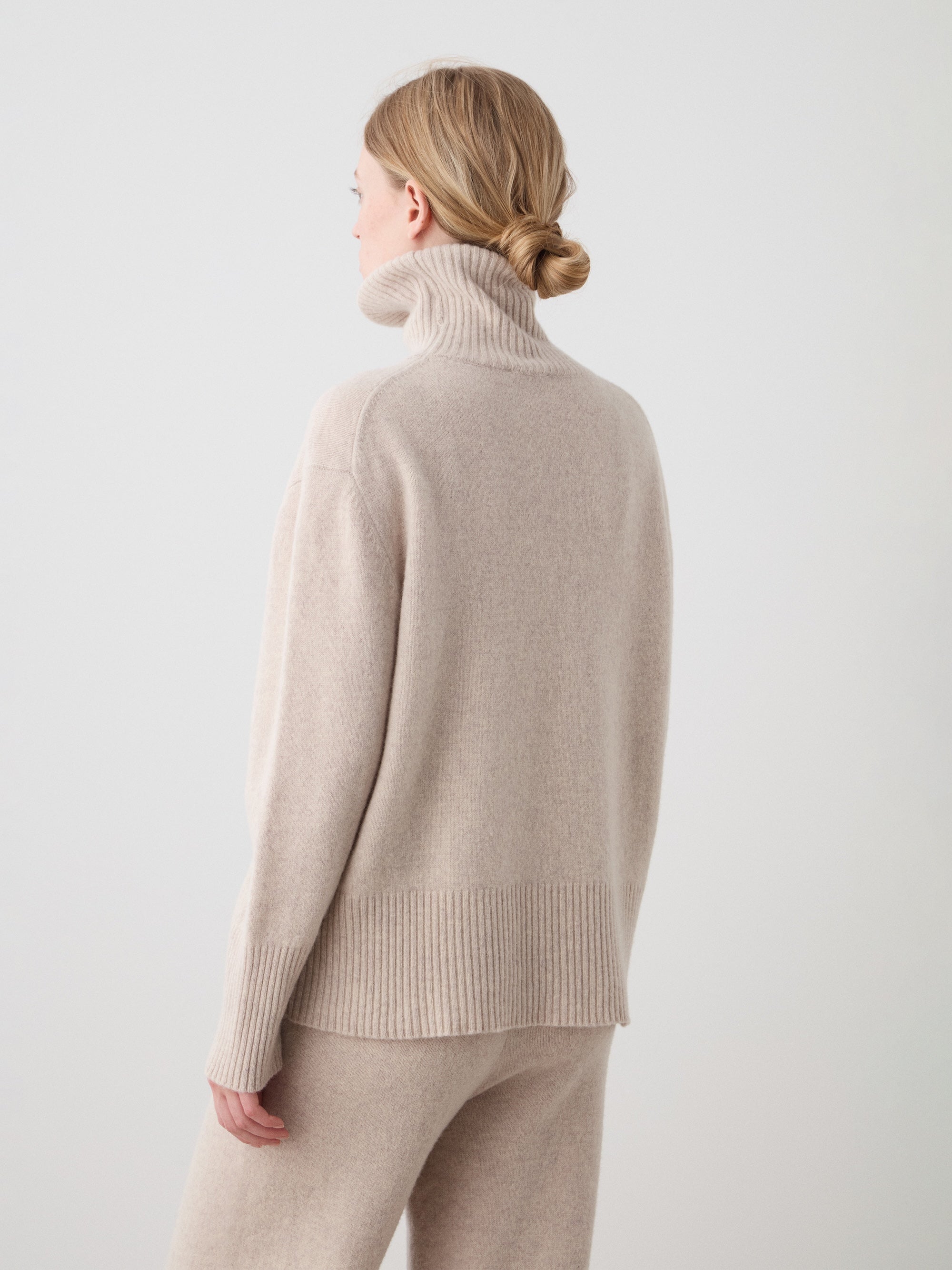 A woman with blonde hair in a low bun wears the We Norwegians Geilo BoyFriend Sweater women, with matching pants, standing with her back to the camera against a plain light background.