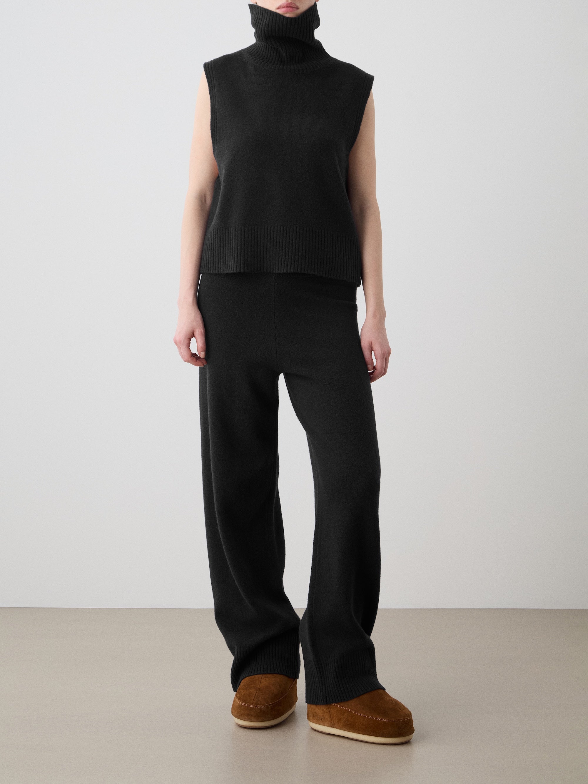 A person stands against a plain background wearing the We Norwegians Geilo Vest women in black extrafine Merino wool, paired with matching loose black knit pants and brown slip-on shoes. Their face is not visible.