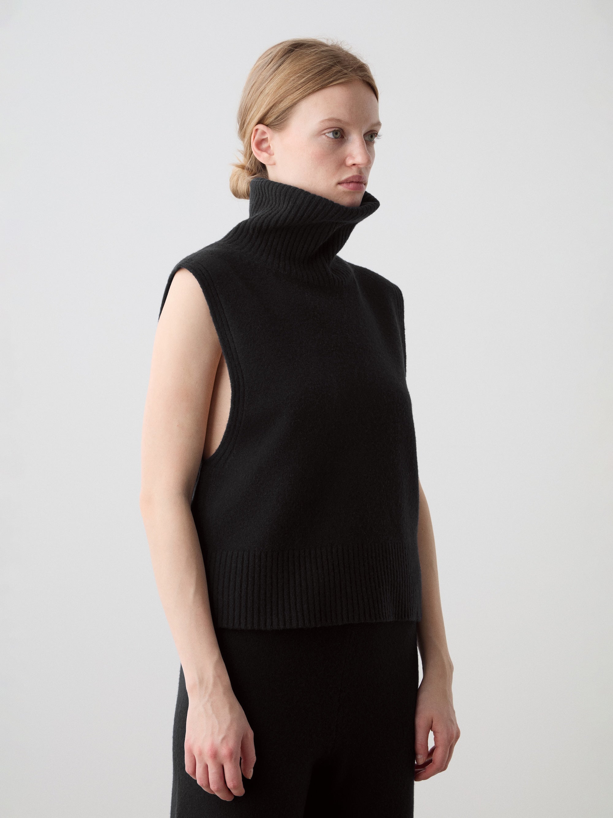 A fair-skinned person with light brown hair in a low bun wears the We Norwegians Geilo Vest women in black, crafted from Extrafine Merino Wool, and black pants, standing against a plain light grey background.