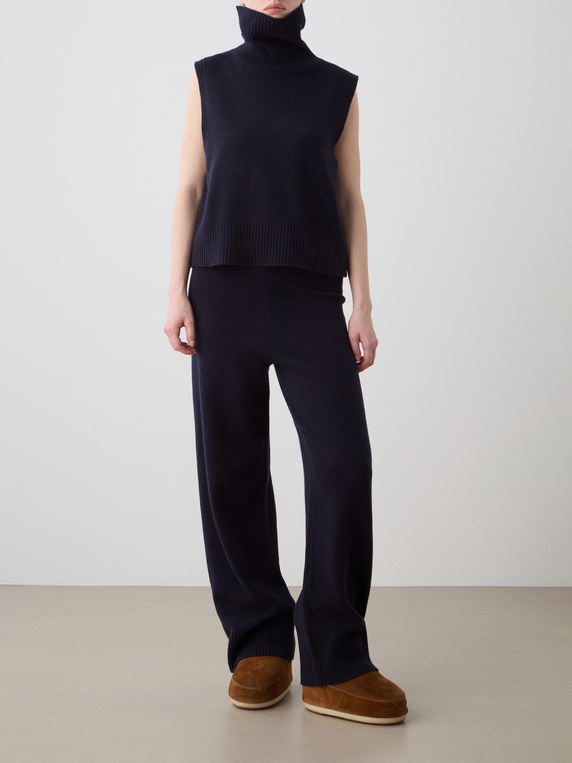A person stands indoors in a We Norwegians Geilo Vest women, made of navy extrafine merino wool, styled with matching wide-leg knit pants and brown slip-on shoes. Their relaxed hands are at their sides, and their face is not shown.