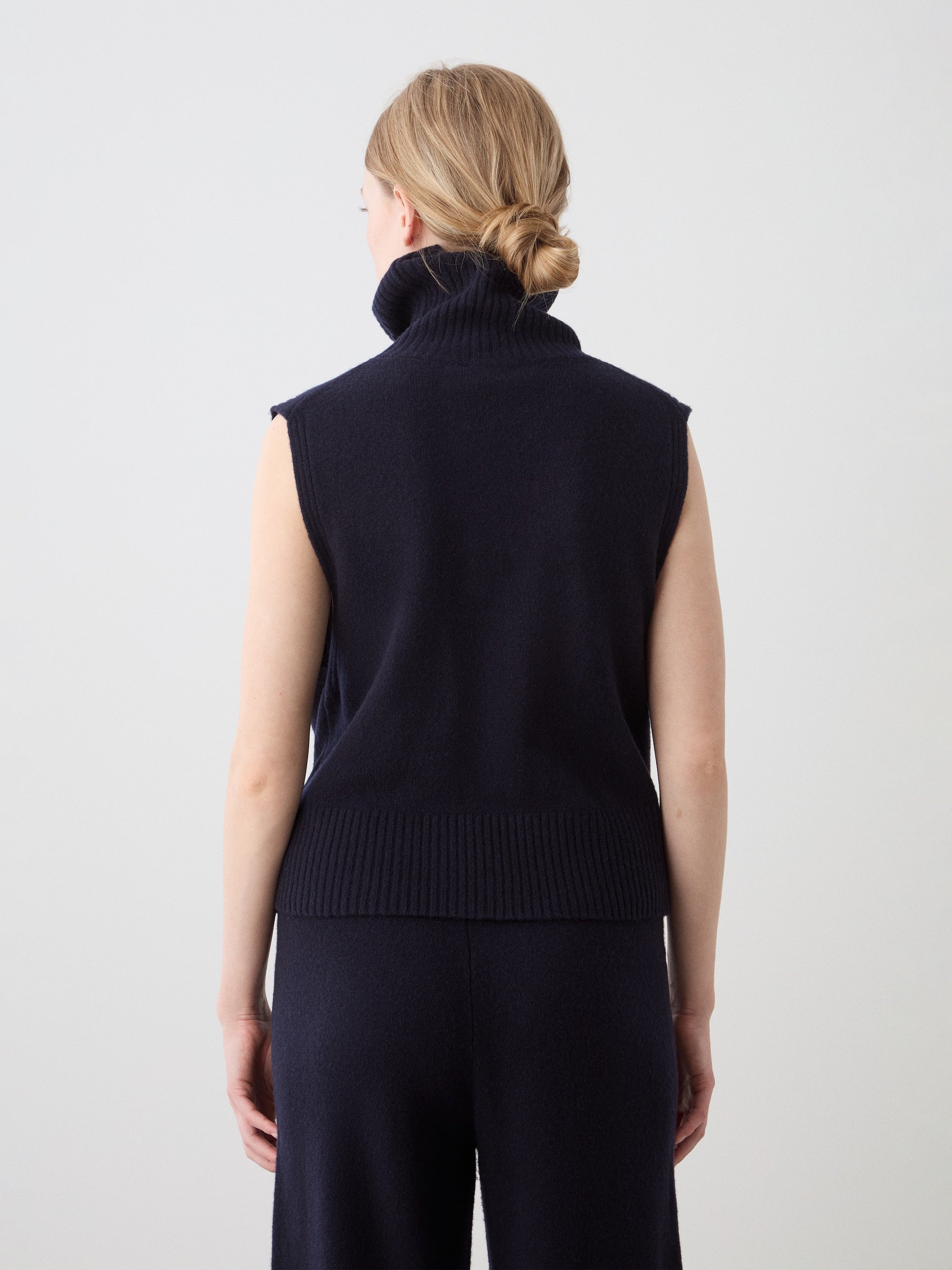 A woman with light skin and blonde hair in a low bun is shown from the back wearing the We Norwegians Geilo Vest women—an extrafine merino wool, sleeveless high-neck dark navy vest and matching pants against a plain background.