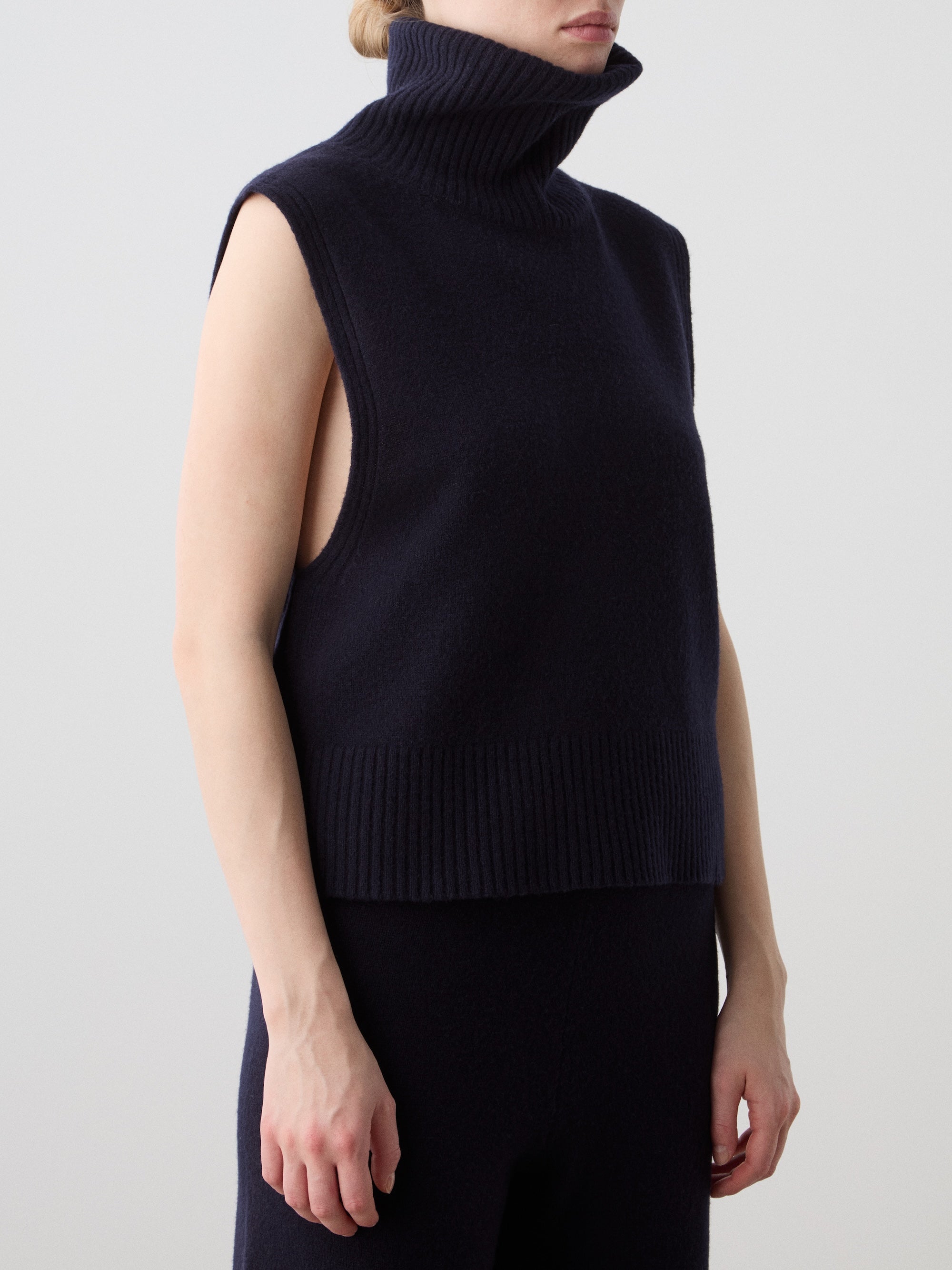 A woman wearing the We Norwegians Geilo Vest—crafted from extrafine merino wool with wide armholes and ribbed hems—stands against a plain light background.