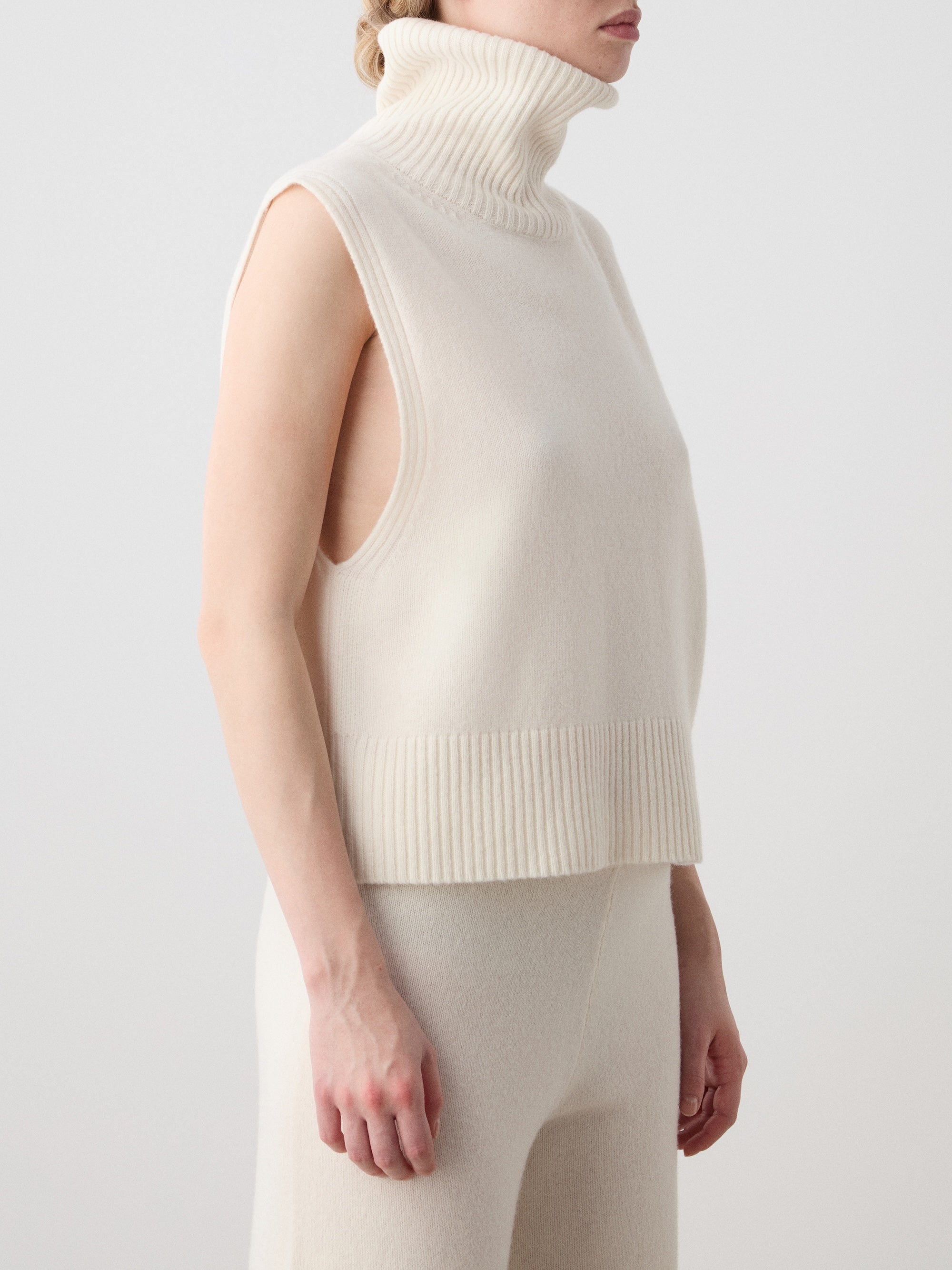 A woman wears the We Norwegians Geilo Vest in off-white, crafted from Extrafine Merino Wool, paired with matching pants. She stands sideways against a plain light background with her face partially out of frame.