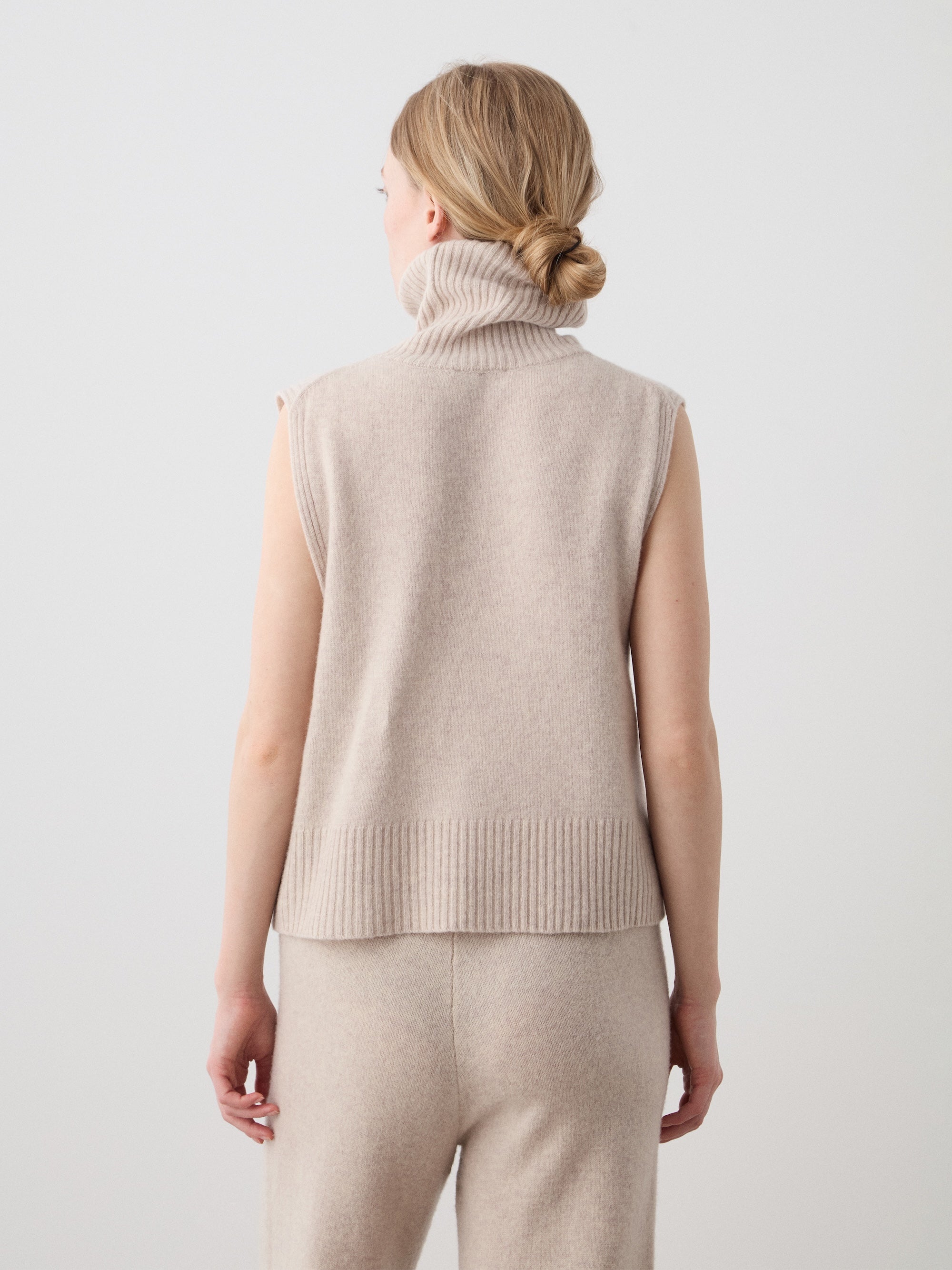 A woman with blonde hair in a low bun is seen from behind wearing the sleeveless beige Geilo Vest from We Norwegians, made of extrafine merino wool, paired with matching pants against a plain light background.
