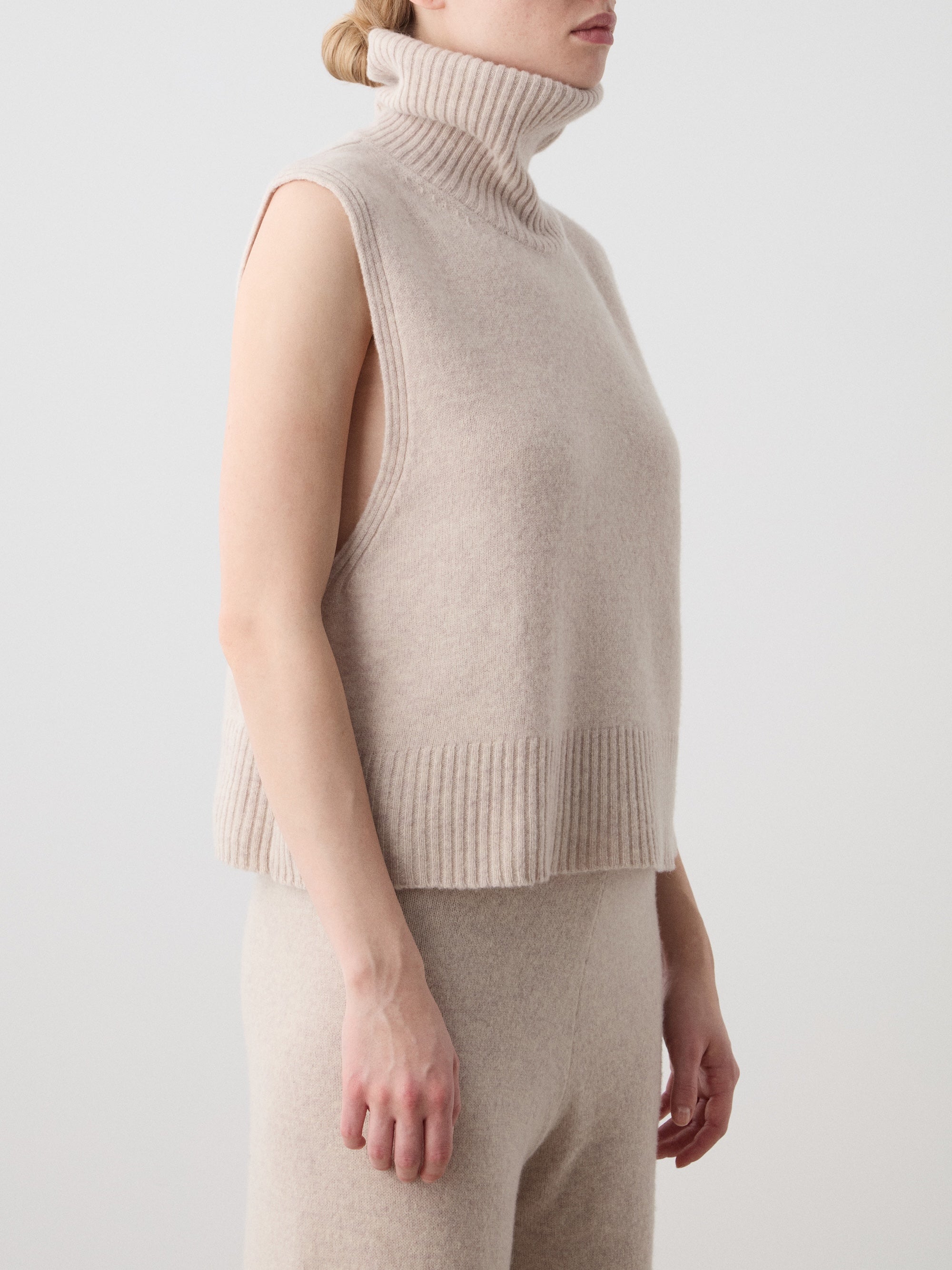 A person wears the We Norwegians Geilo Vest women in sleeveless light beige extrafine merino wool with matching high-waisted pants, standing against a plain light background. Their face is partially out of frame.