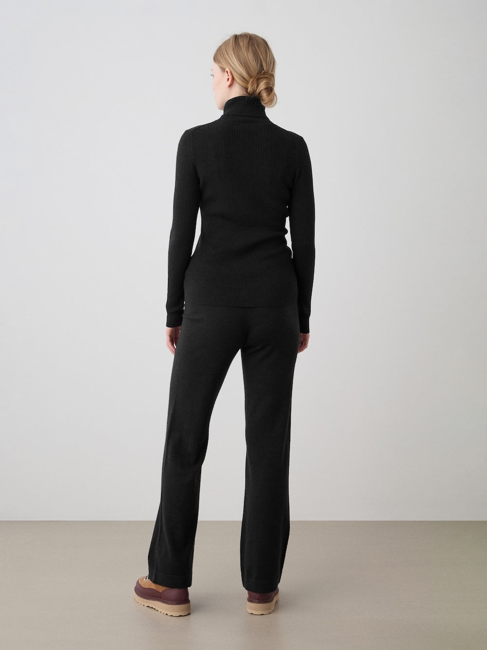 A person with blond hair in a low bun stands facing away, wearing the We Norwegians Geilo TurtleNeck for women with black pants and brown shoes, set against a plain light-colored wall and floor.