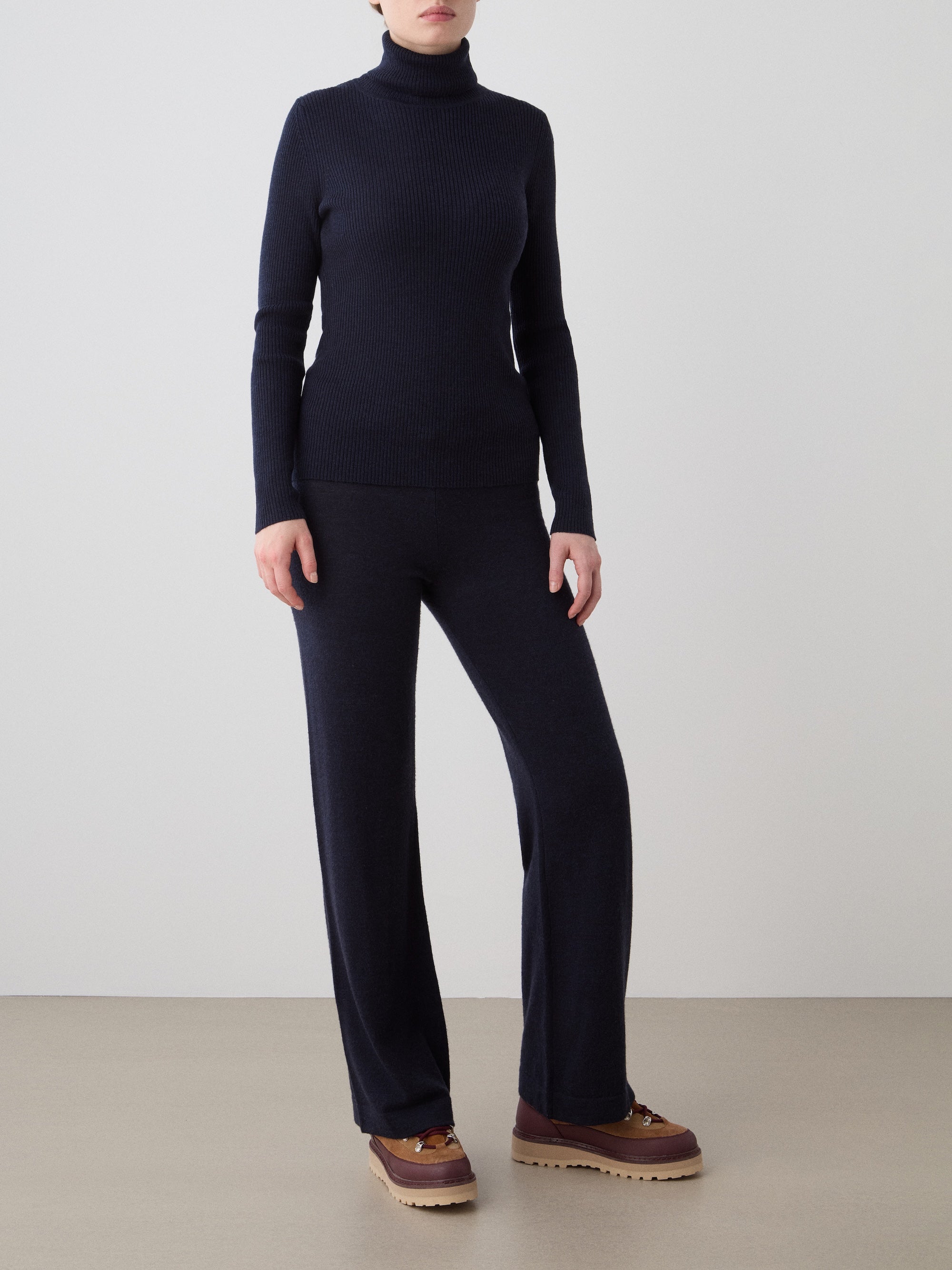 A person stands against a plain background wearing the We Norwegians Geilo Turtleneck for women with matching wide-legged pants and brown platform shoes. Their hands are relaxed by their sides, and their face is not visible.