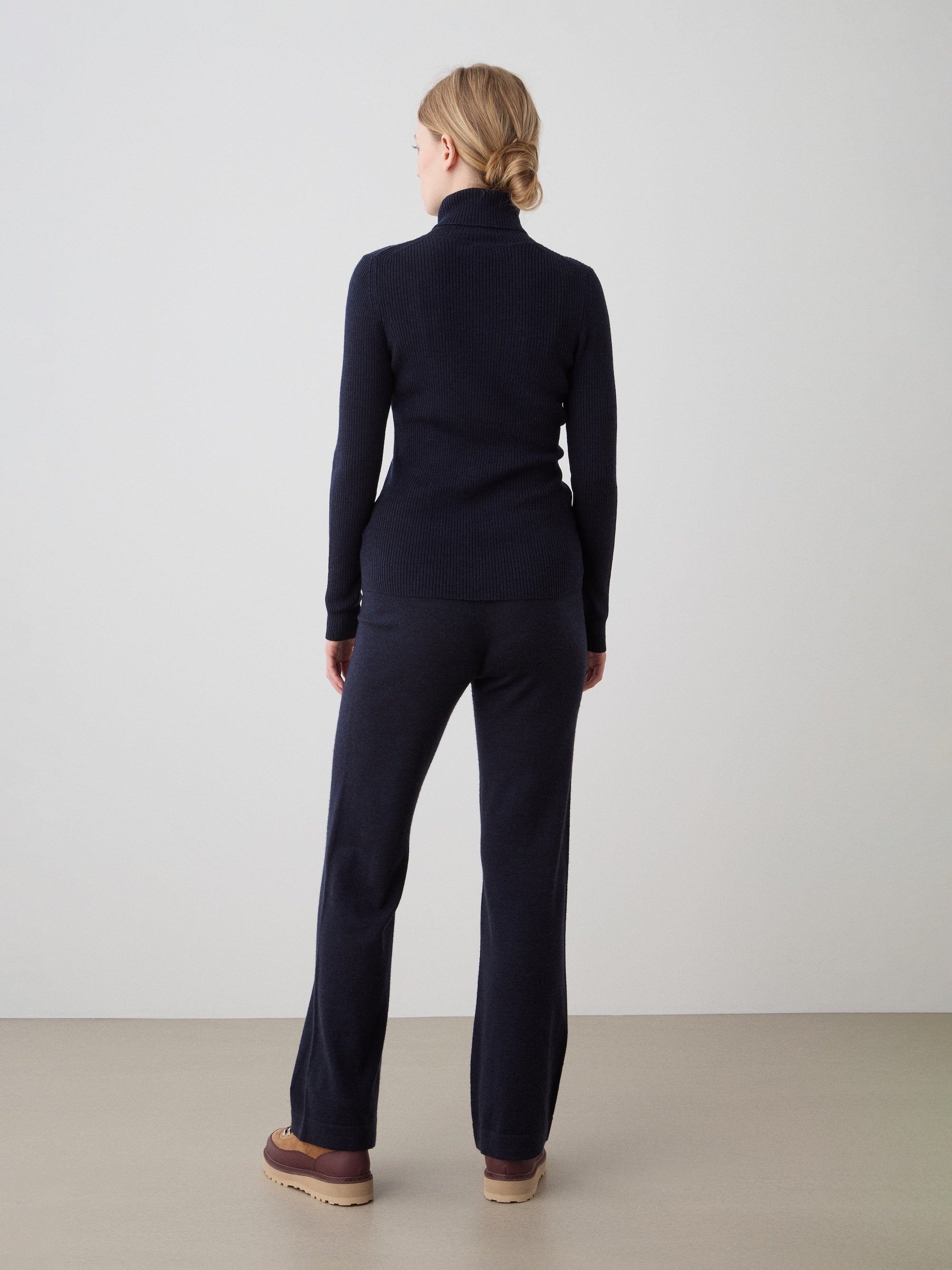A woman with blonde hair in a low bun stands facing away from the camera, wearing the We Norwegians Geilo TurtleNeck for women in navy, paired with matching pants and brown platform shoes against a plain light background.