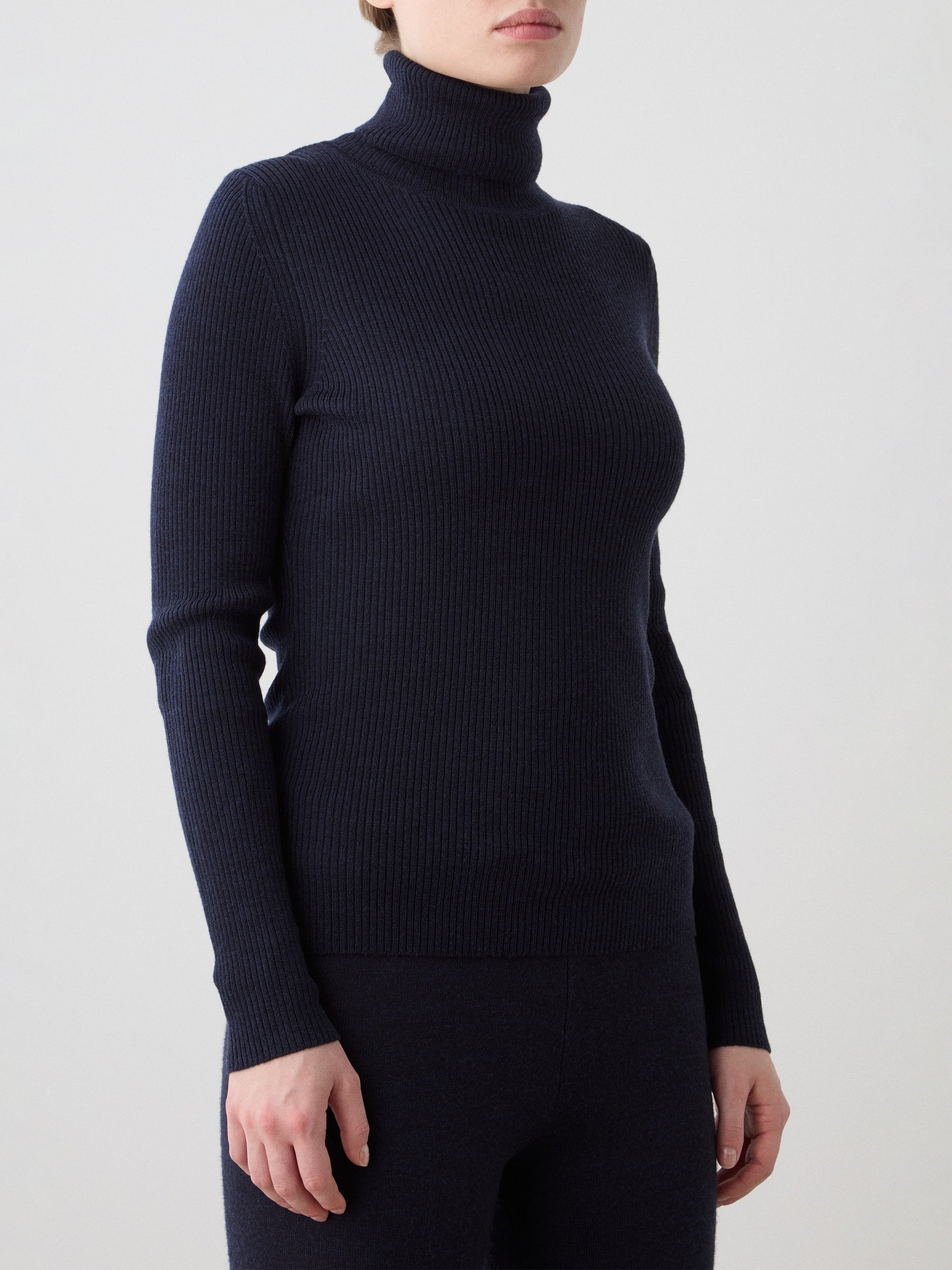 A person wears the We Norwegians Geilo TurtleNeck women, a fitted dark navy ribbed top with a roll neck collar, paired with matching pants, standing against a plain light background with their head partially out of frame.