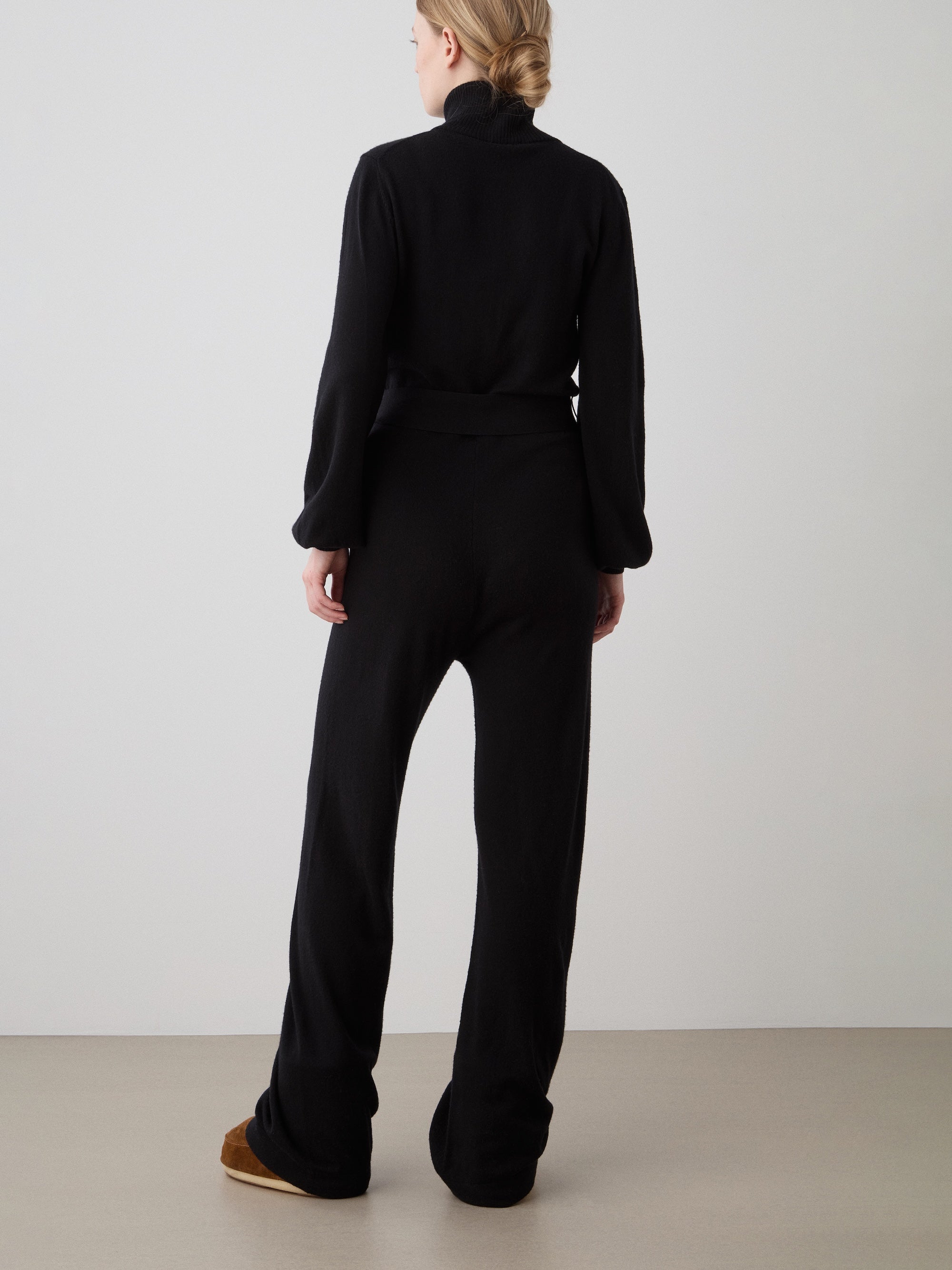 A woman with blonde hair tied back stands facing away, wearing the We Norwegians Geilo Jumpsuit for women and brown shoes—ideal for a stylish mountain resort look—set against a plain light background.
