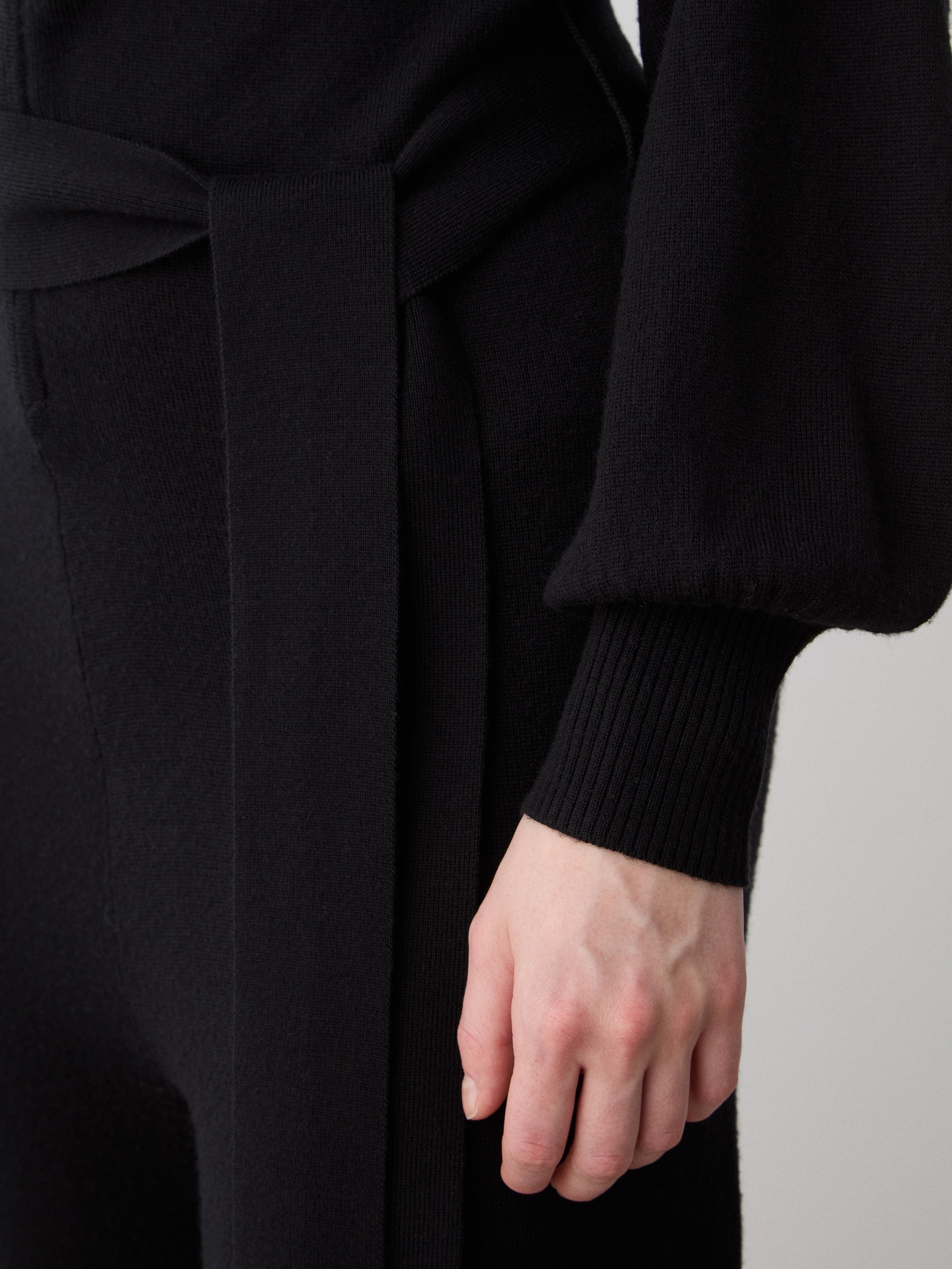 A woman wears the We Norwegians Geilo Jumpsuit—black, long-sleeved with a matching fabric belt—her right hand relaxed at her side against a neutral background.