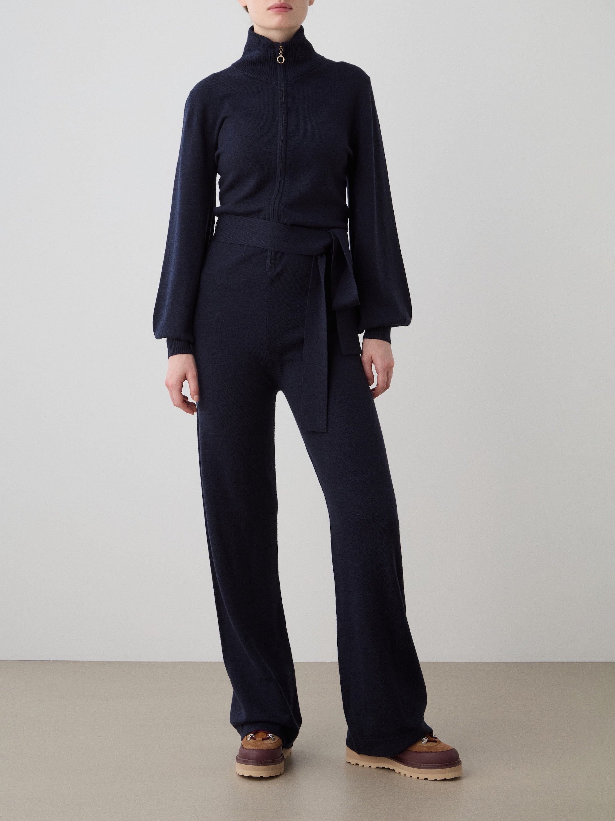 A woman models the We Norwegians Geilo Jumpsuit in dark navy, featuring long sleeves, a high zip-up collar, waist tie, and wide-leg pants. She pairs it with tan platform shoes against a plain light background.