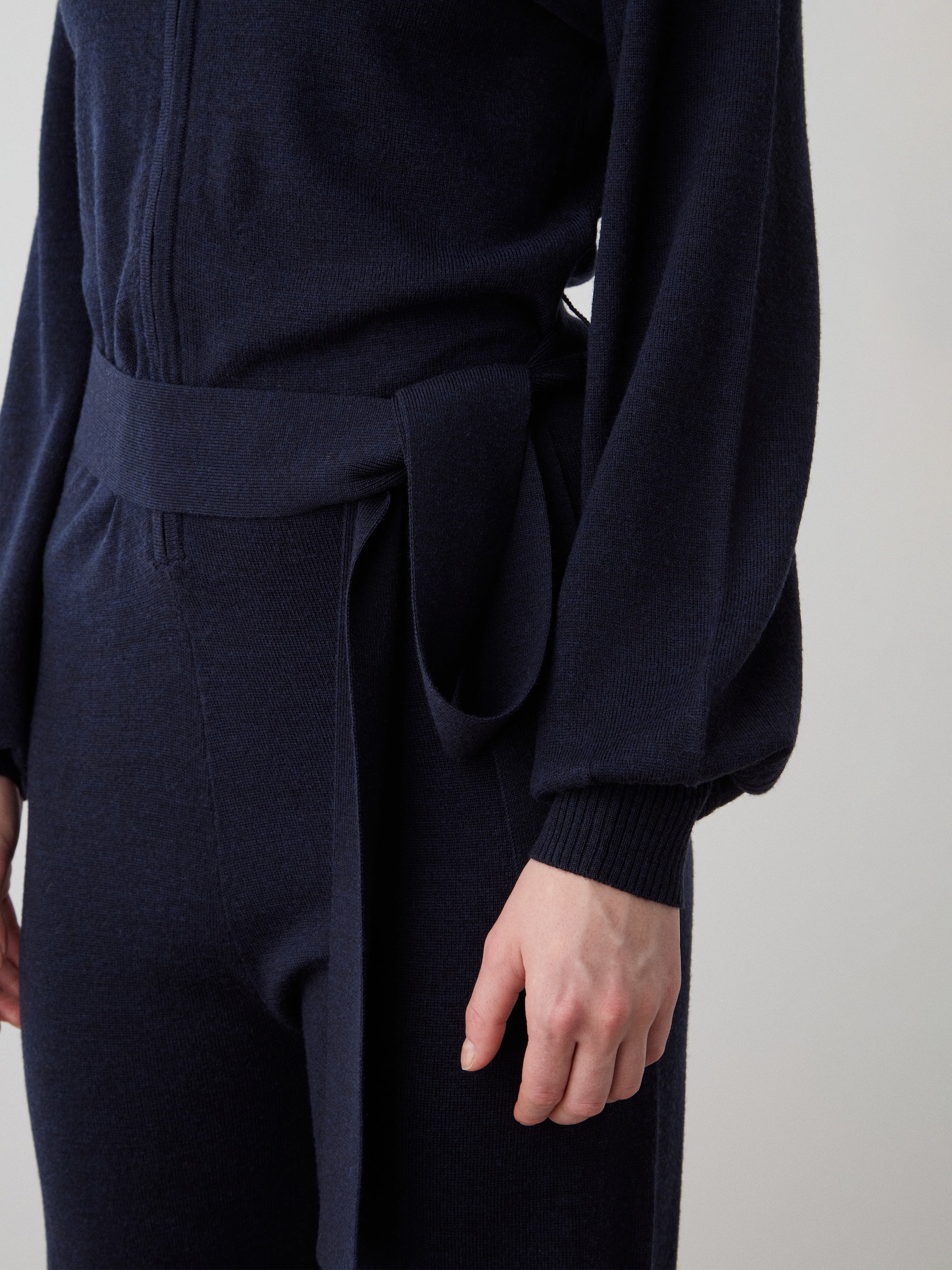 A model showcases the We Norwegians Geilo Jumpsuit for women in dark navy, featuring long sleeves and a fabric belt at the waist, photographed from shoulder to mid-thigh against a plain light background.