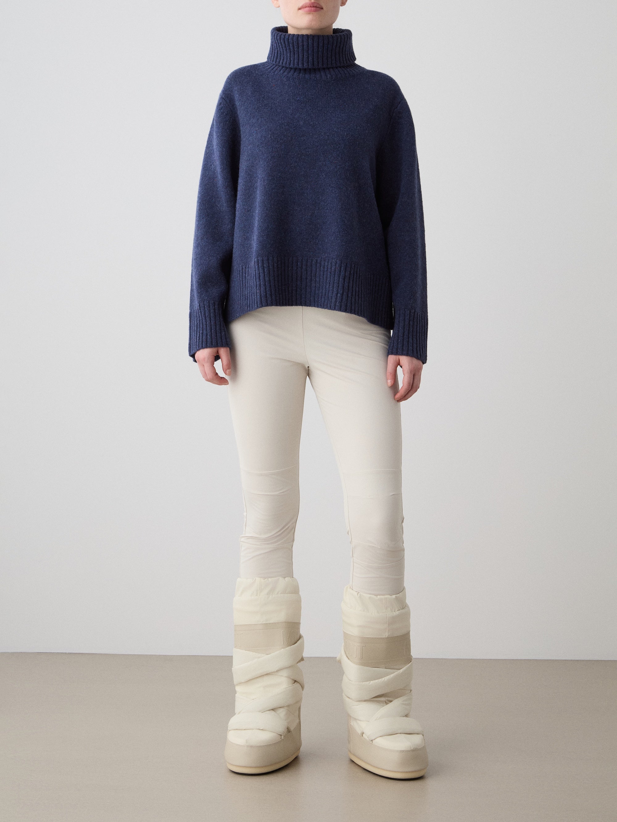A person stands indoors in a navy blue Highland Sweater women by We Norwegians, made from Extra Fine Merino Wool, paired with cream fitted pants and off-white padded winter boots against a plain background.
