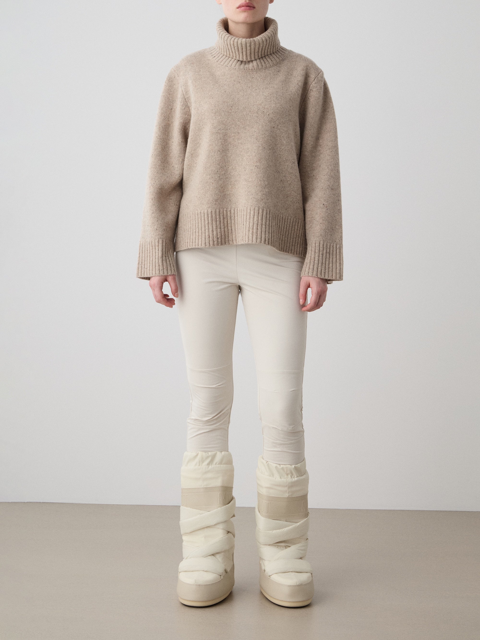 A person, head not visible, stands against a plain background wearing the beige Highland Sweater women by We Norwegians in Extra Fine Merino Wool, paired with cream leggings and beige puffy winter boots.