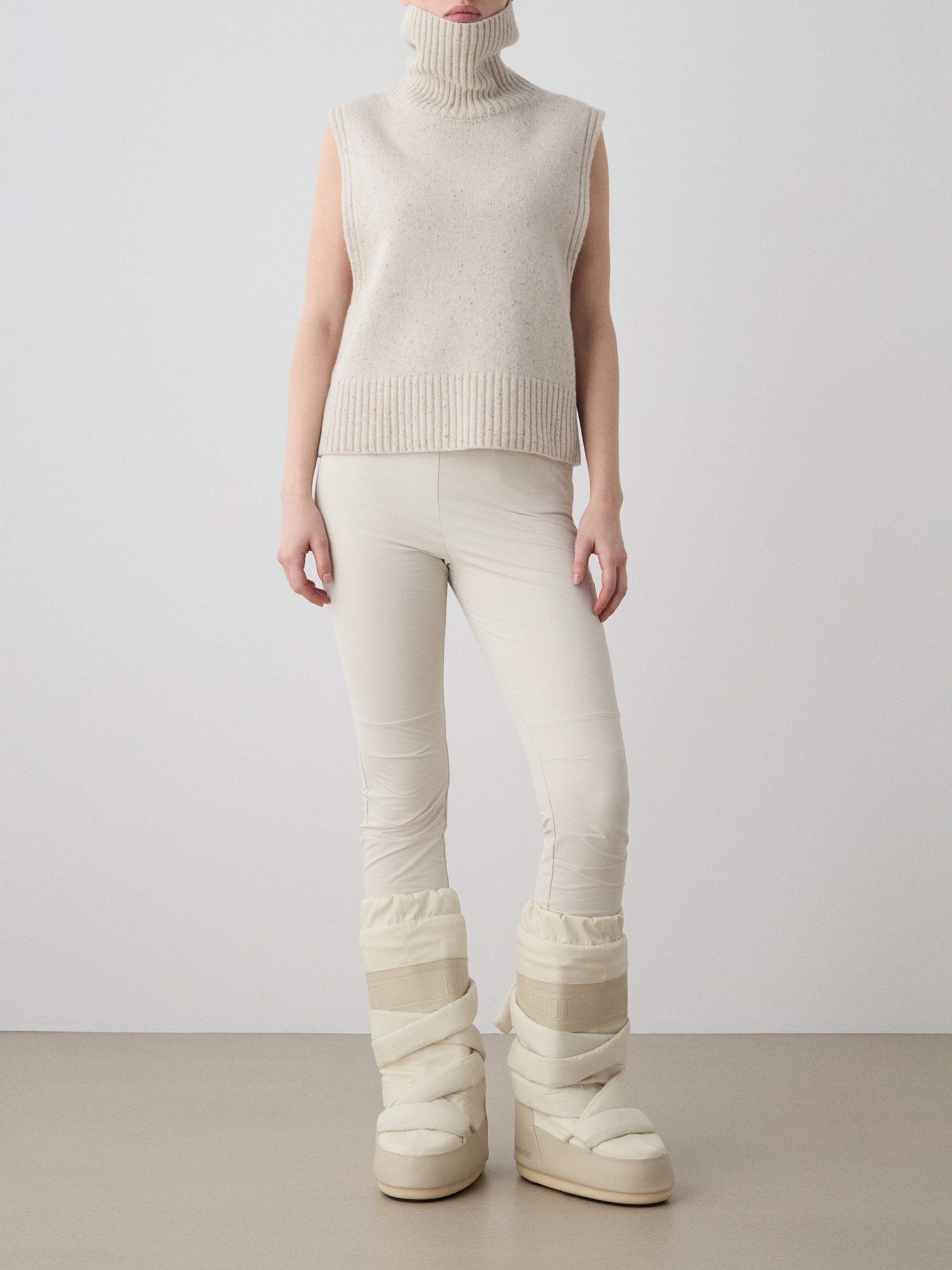 A person stands against a plain background wearing the We Norwegians Highland Vest for women, fitted pants, and large padded boots—all in beige and off-white tones.