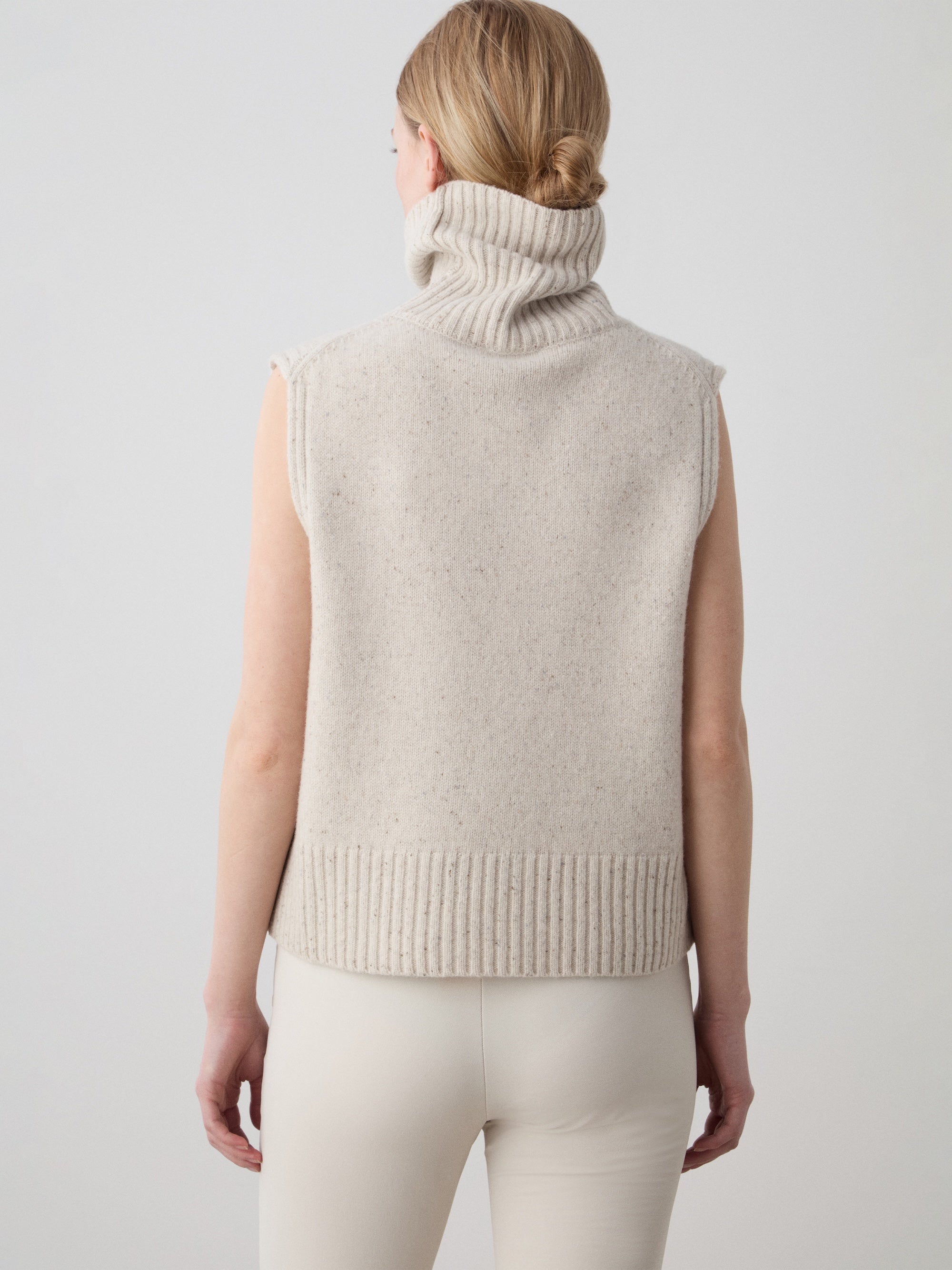 A woman with blonde hair in a low bun is shown from the back, wearing the We Norwegians Highland Vest for women—a sleeveless, light beige layer—paired with cream pants against a plain light background.