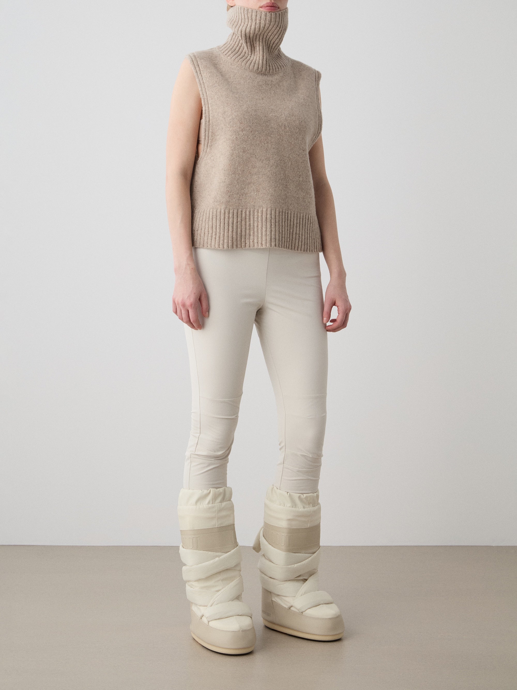 A person wears the We Norwegians Highland Vest for women, styled with fitted ivory pants and chunky cream-colored padded boots. The merino wool turtleneck partially covers their face against a plain background.