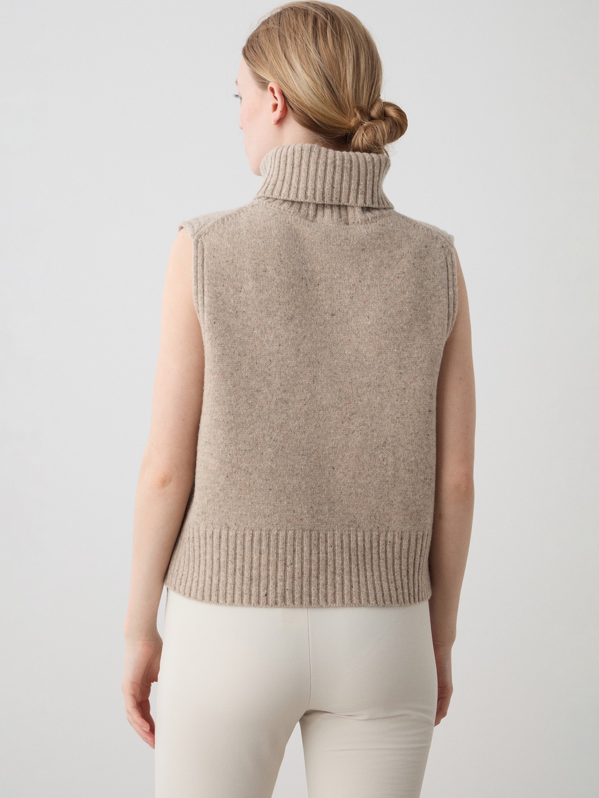 A woman with light brown hair in a low ponytail wears the We Norwegians Highland Vest for women and light pants, standing with her back to the camera against a plain light background.