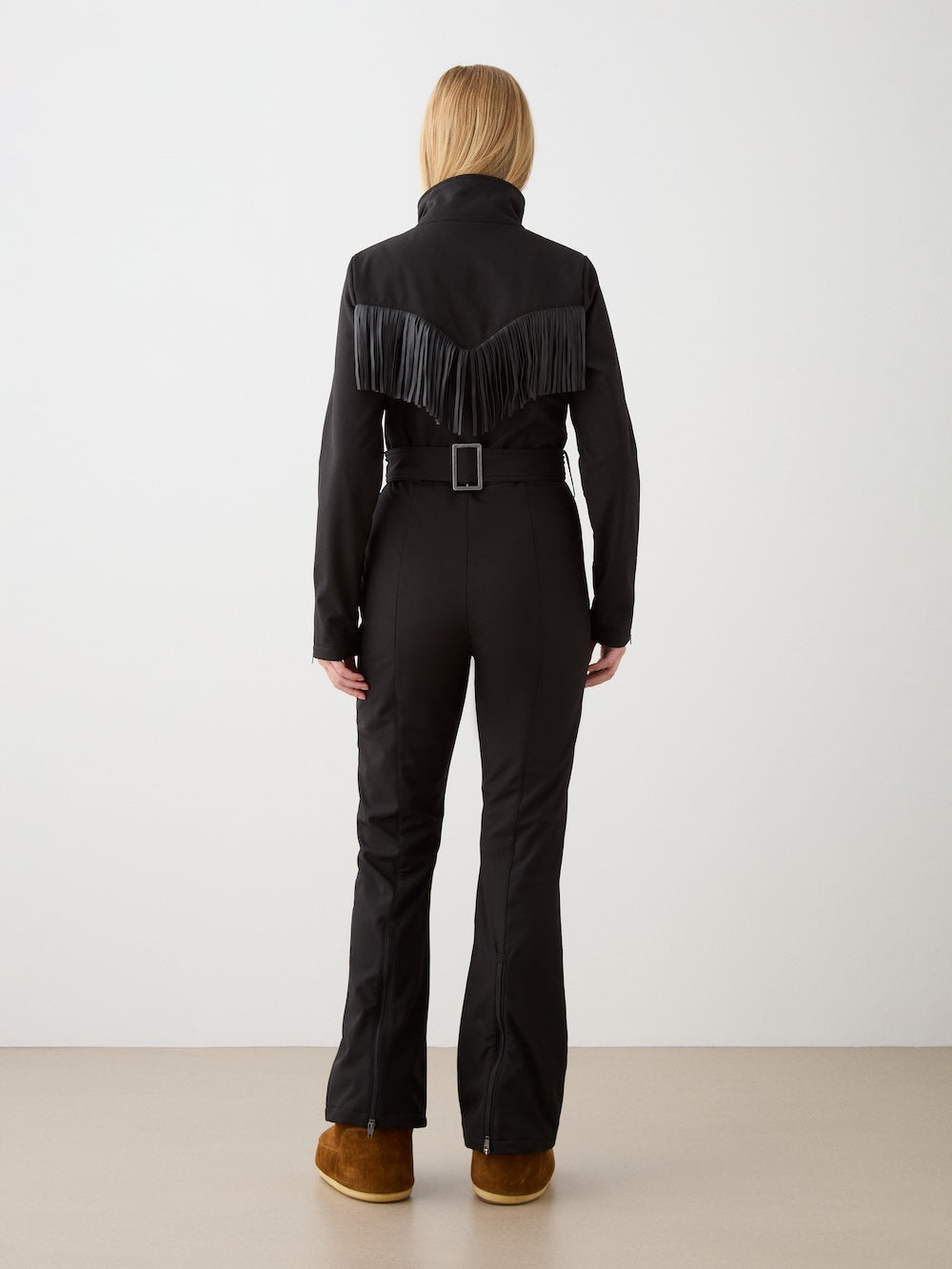A person faces away, wearing the We Norwegians Western Ski Suit for women—a fitted black suit with a belted waist and fringe across the back—paired with brown boots, against a plain light background.