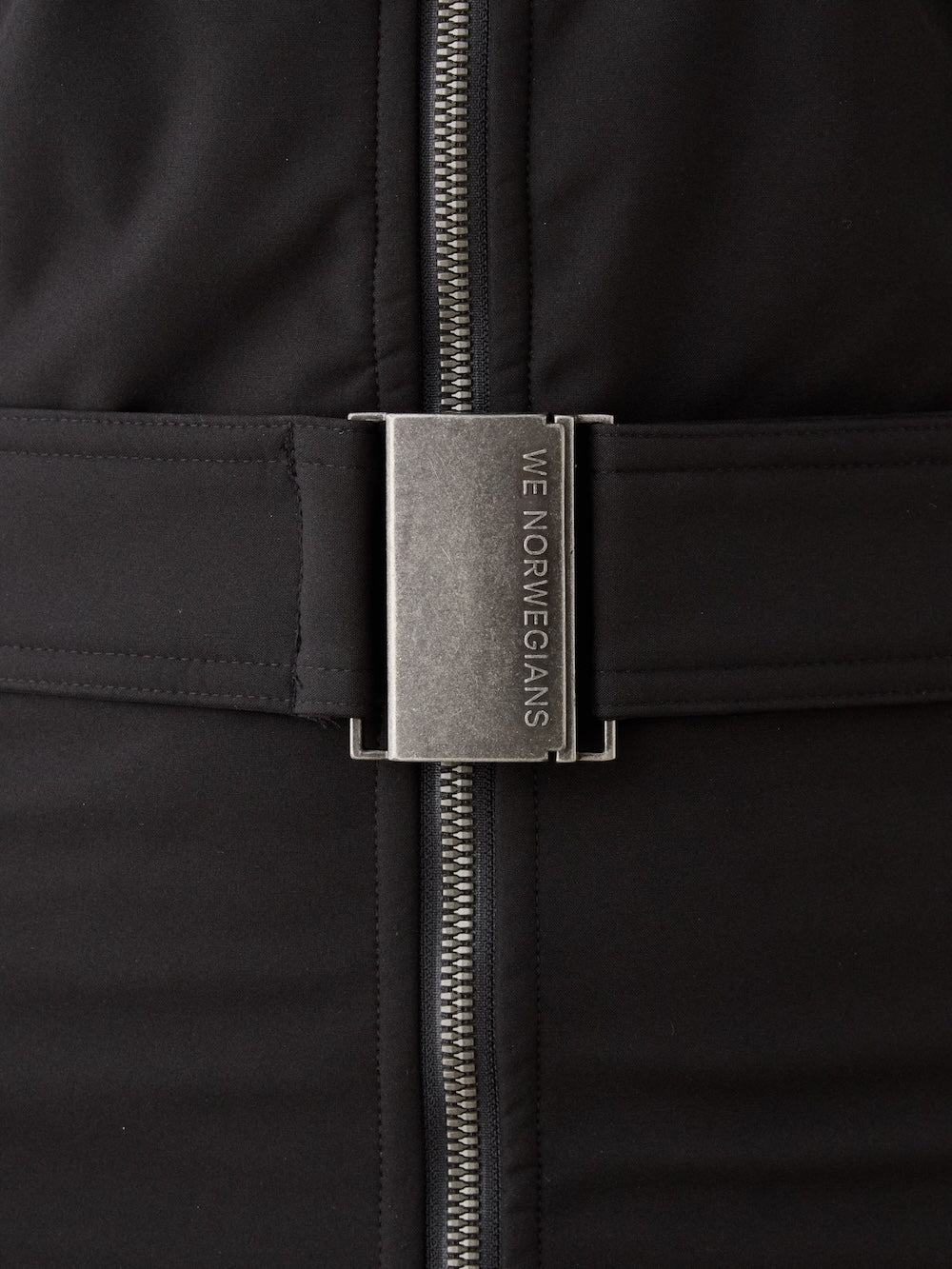 Close-up of the Western Ski Suit for women by We Norwegians, showcasing a sleek black design with a vertical silver zipper and metallic belt buckle engraved "WE NORWEGIANS," highlighting sophisticated après-ski style.