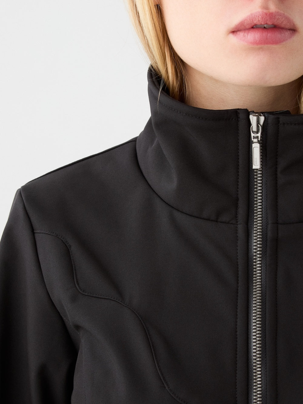 Close-up of a person in the We Norwegians Western Ski Suit for women, showcasing its high collar and silver zipper. Light hair peeks out, embodying après-ski style and winter sophistication.
