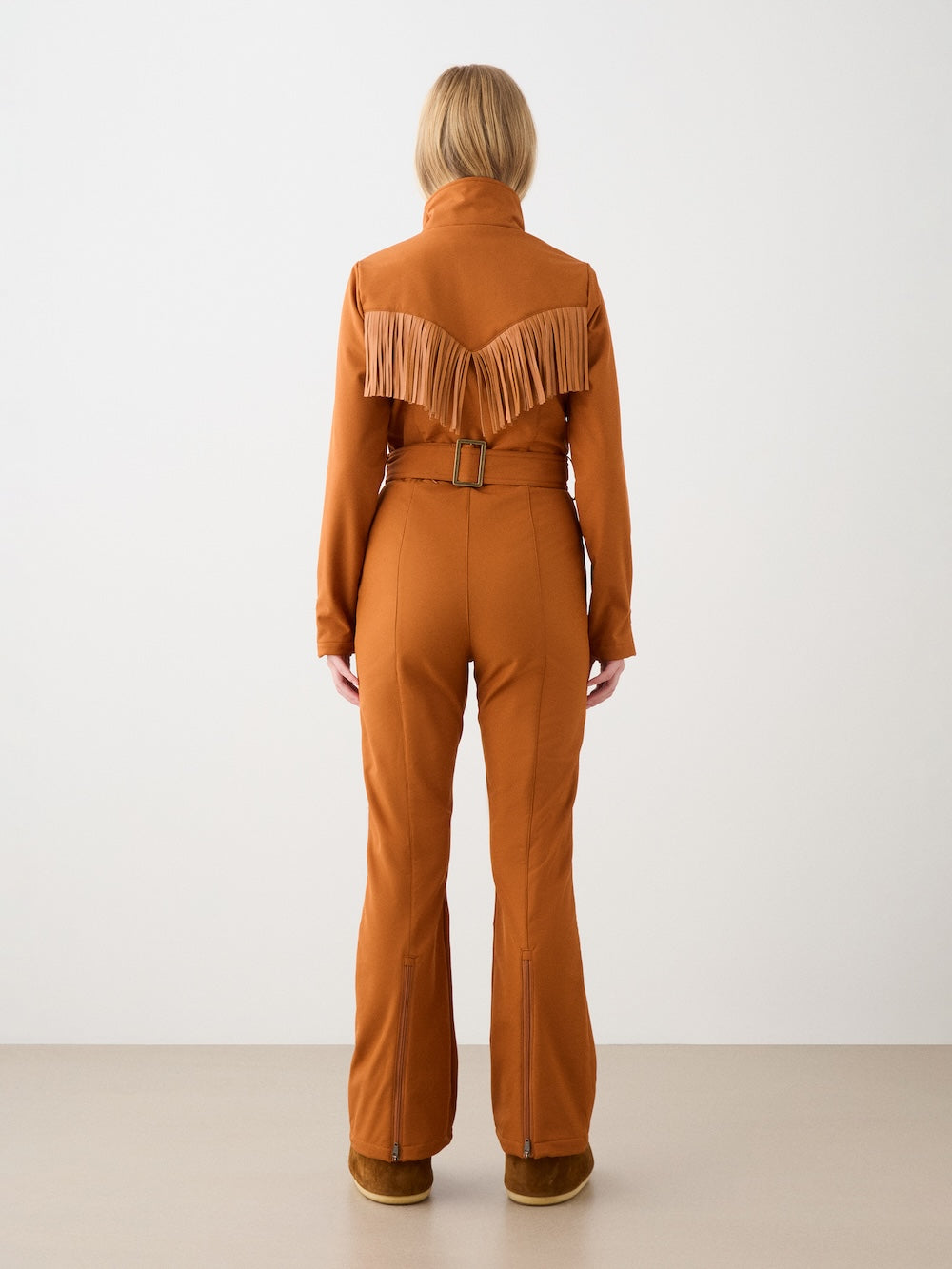 A person with short blonde hair wears the We Norwegians Western Ski Suit for women, featuring a rust color, fringe-accented back, belted waist, flared legs, and long sleeves. The retro-inspired look stands out against a plain light background.