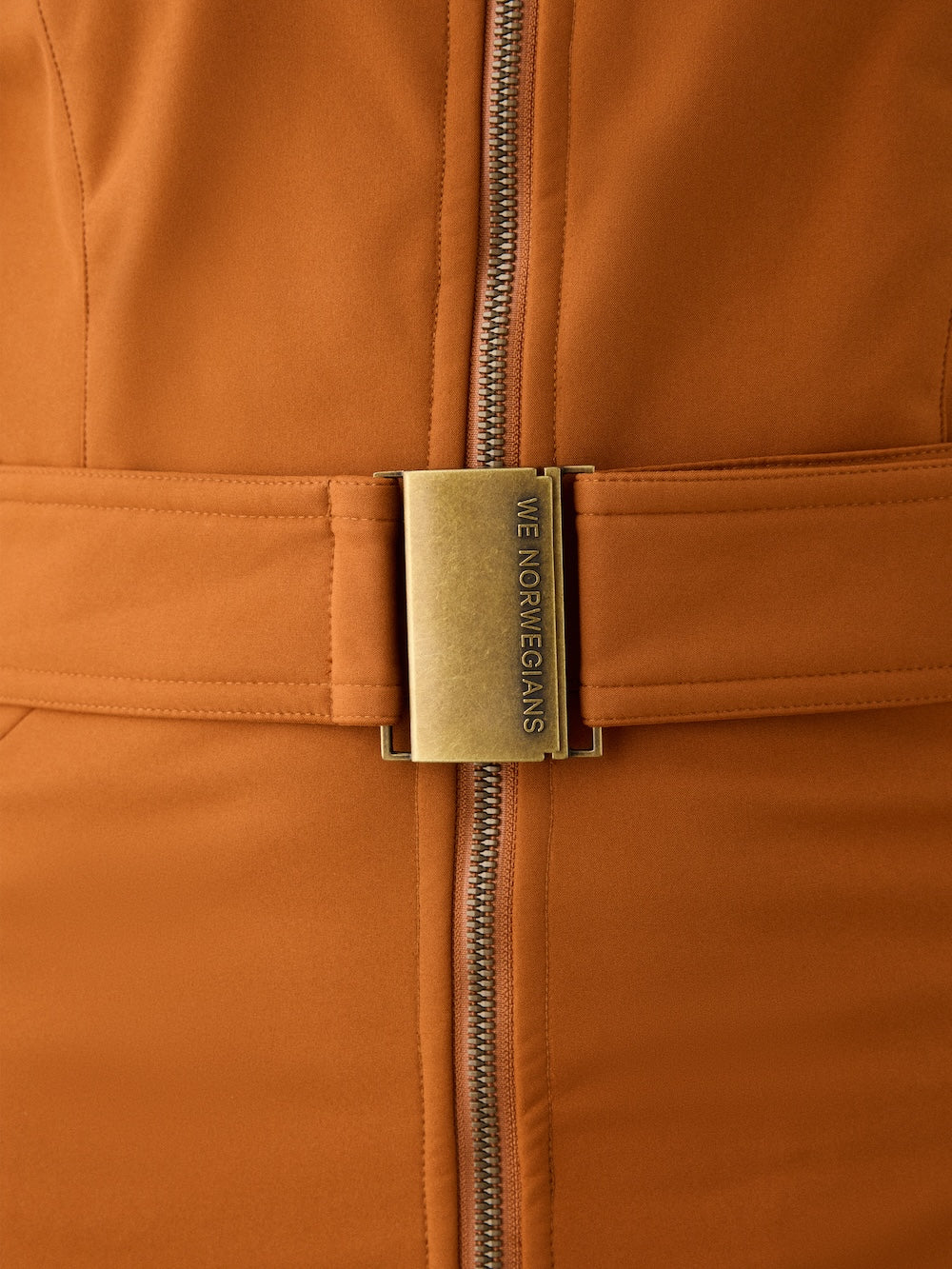 Close-up of the Western Ski Suit for women by We Norwegians, showcasing an orange design with a central zipper and wide belt featuring a gold "WE NORWEGIANS" buckle—ideal for après-ski style.