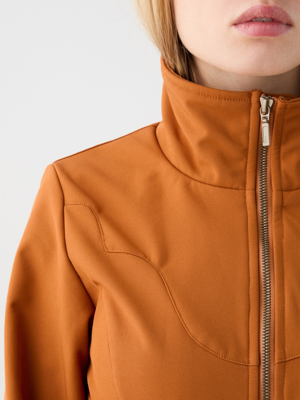 Close-up of a person wearing the We Norwegians Western Ski Suit for women in orange-brown, featuring a high collar and front zipper. The lower face appears against a plain light background, showcasing modern après-ski style.