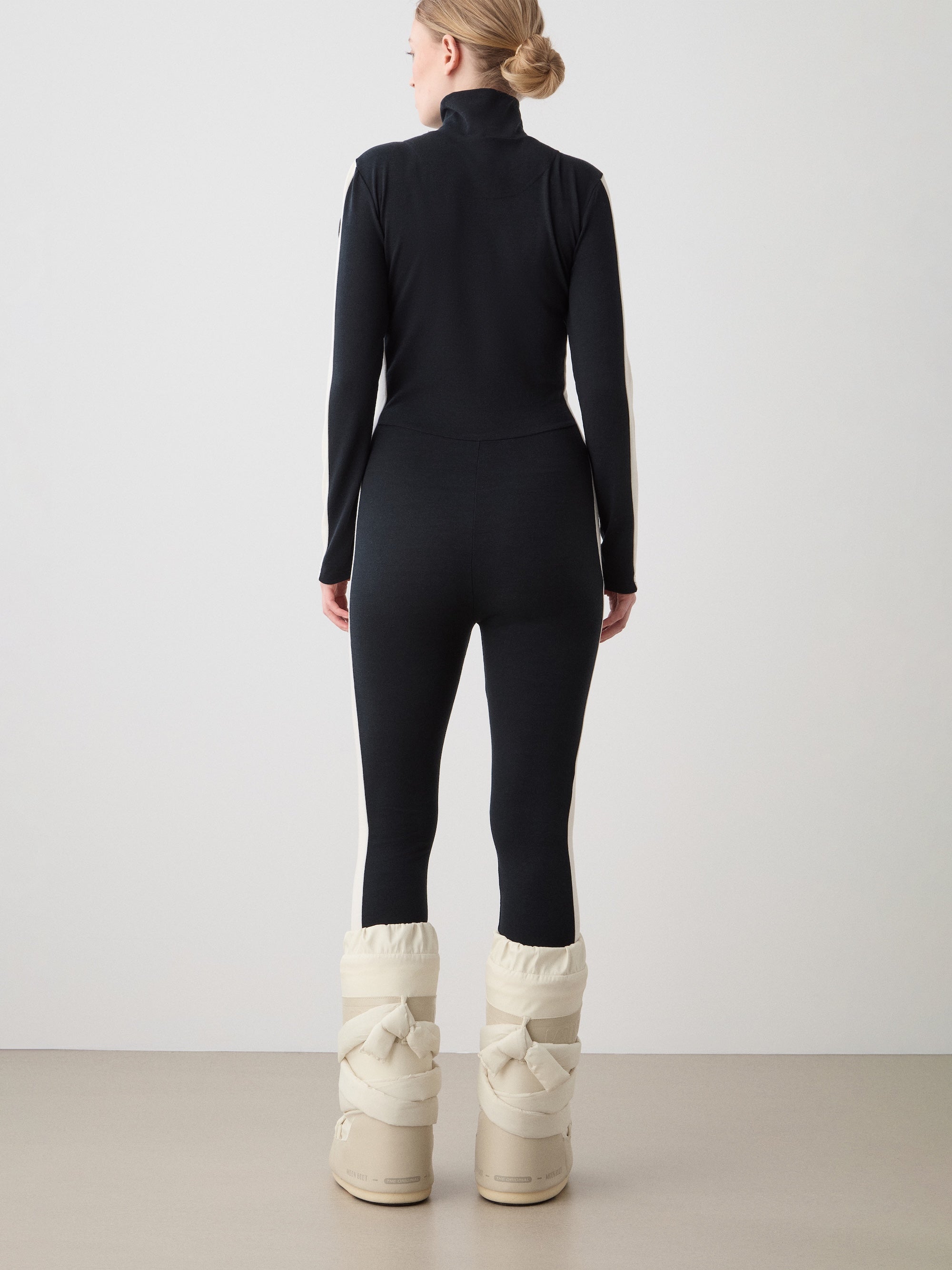 A woman faces away from the camera wearing the We Norwegians Voss Jumpsuit—a sleek black merino piece—and tall, padded off-white winter boots, ideal as a stylish ski base layer, set against a plain light background.
