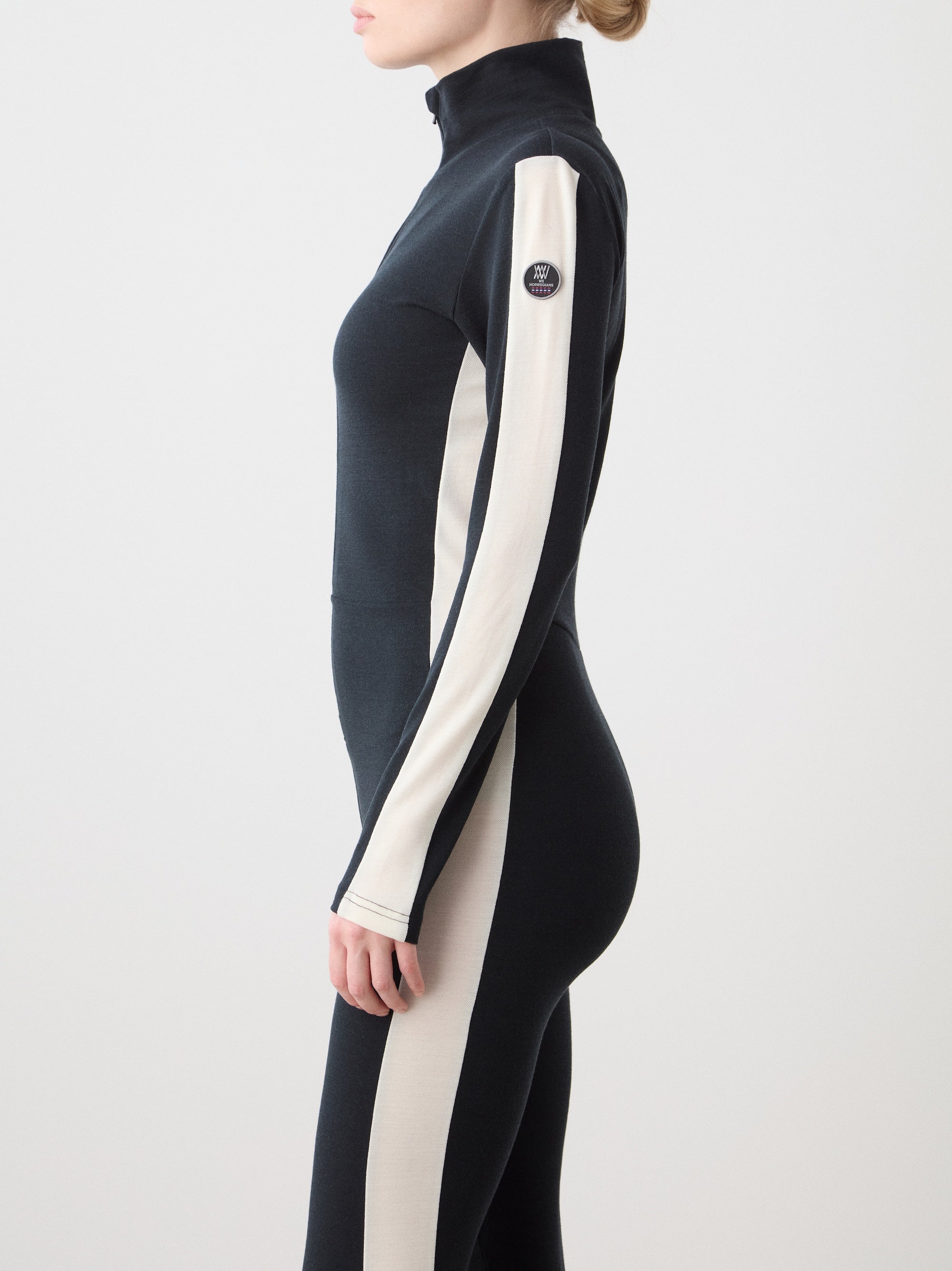 A woman stands in profile against a plain light background wearing the We Norwegians Voss Jumpsuit—a fitted black and cream retro style with a high collar and contrasting cream panels on the side and sleeve.