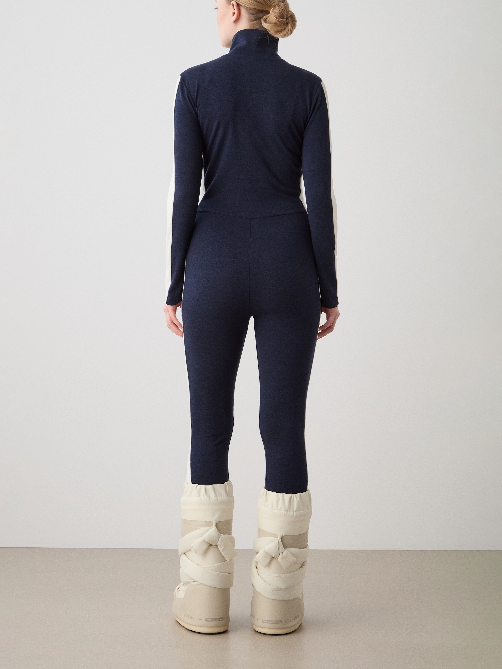 A person stands facing away in the We Norwegians Voss Jumpsuit for women, featuring a navy retro style with cream arm panels and padded winter boots, ideal for ski loungewear, set against a plain light background.