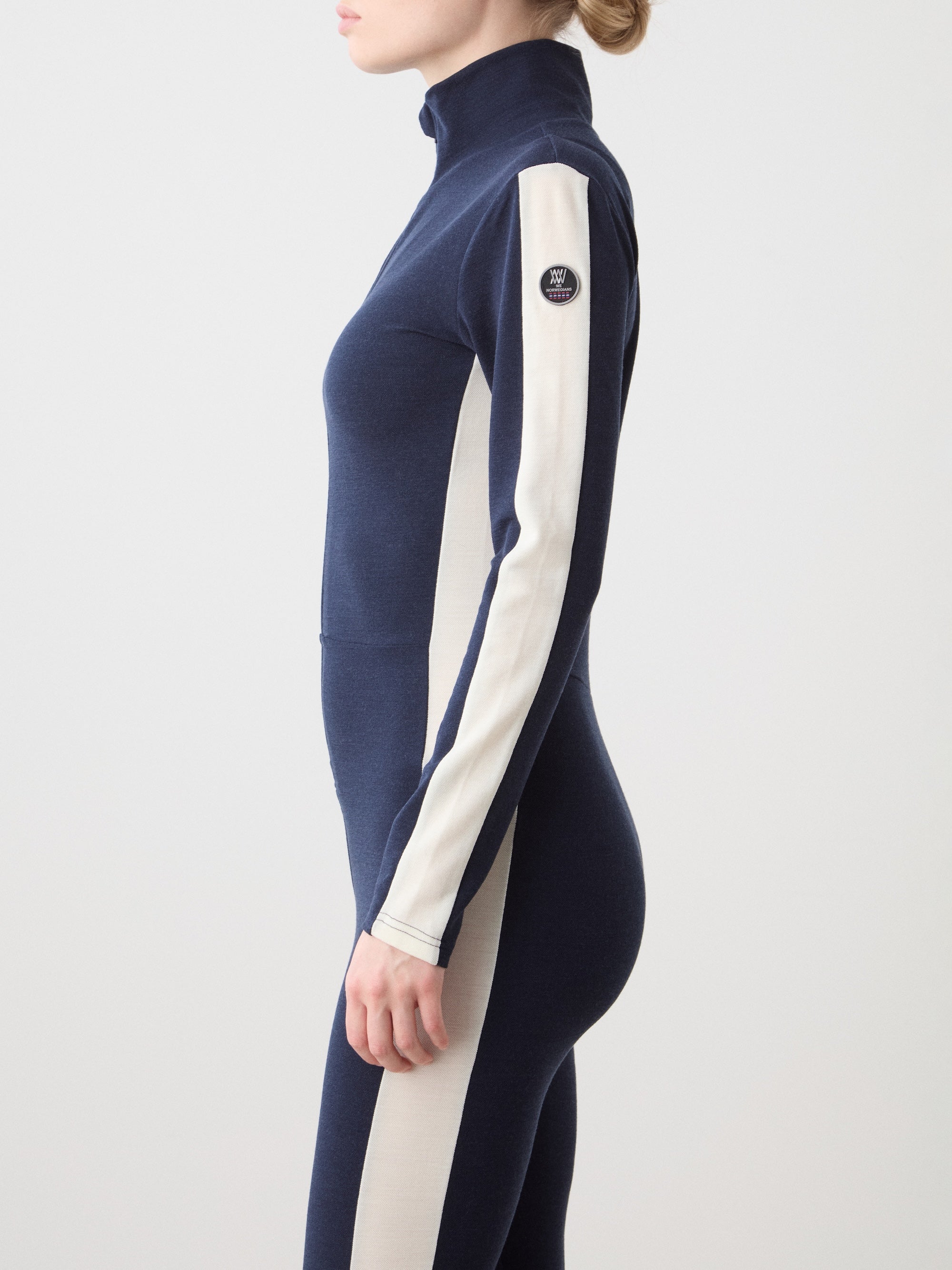 A woman stands sideways against a plain light background, wearing the Voss Jumpsuit by We Norwegians in navy blue with white side panels, a high collar, and a patch logo on the upper arm.