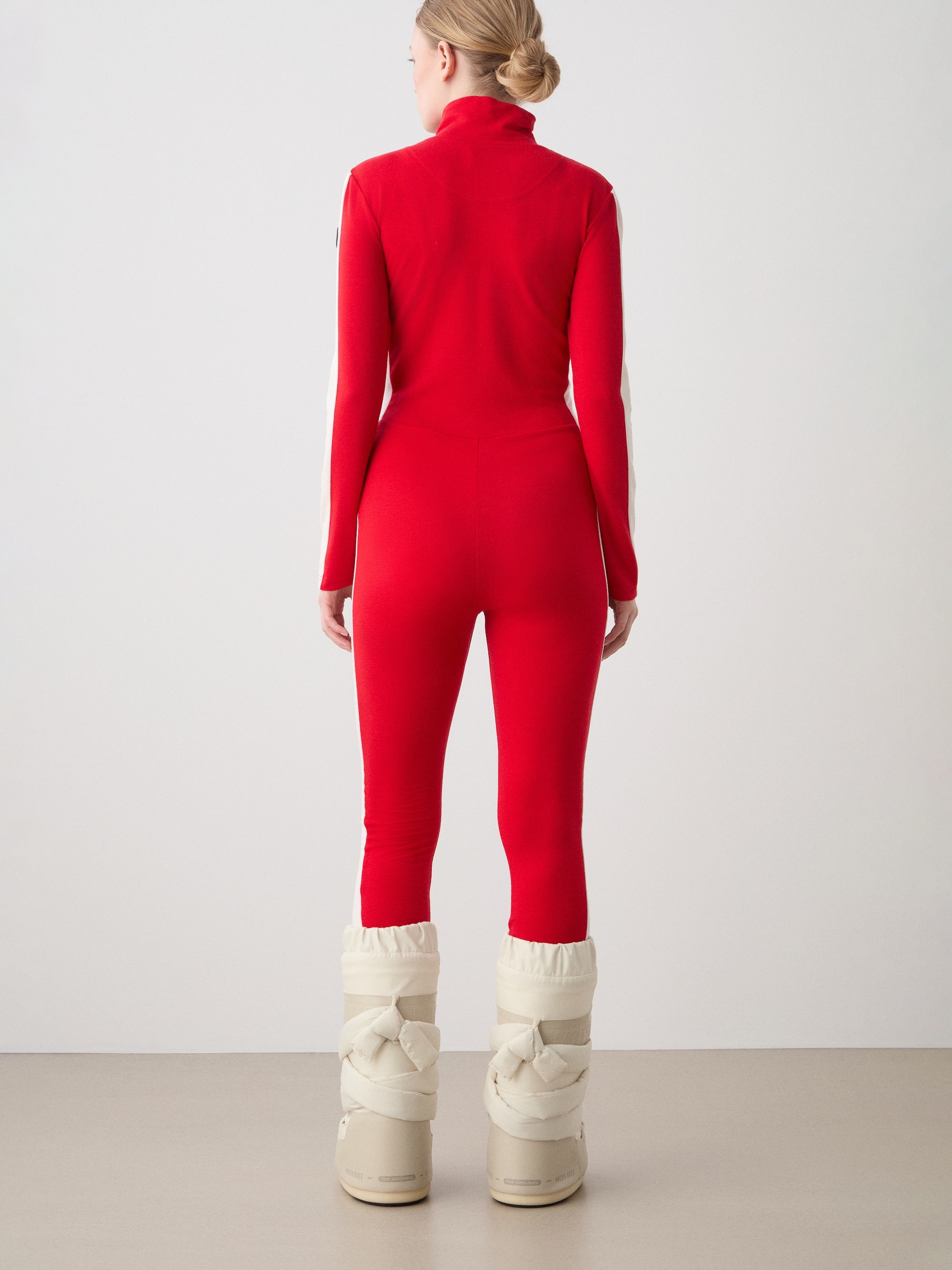 A person stands facing away in the We Norwegians Voss Jumpsuit for women, a fitted red merino one-piece, paired with tall padded white snow boots, against a plain light background.