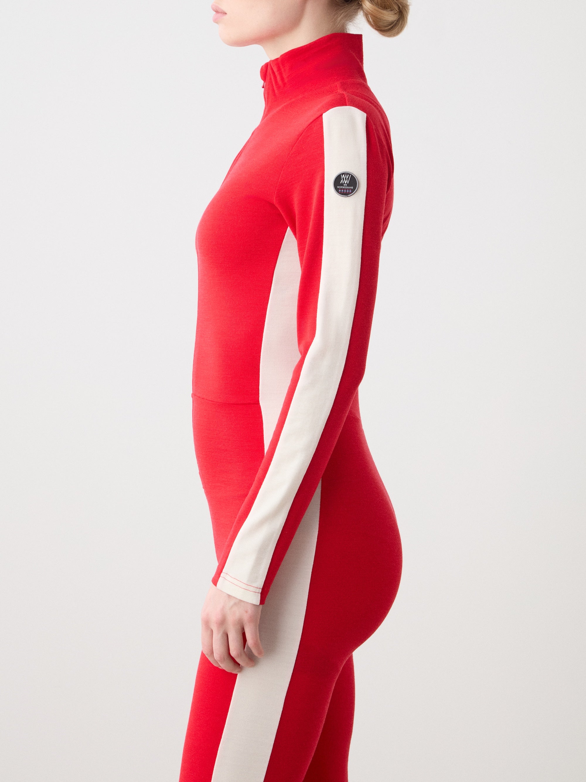 A woman wearing the We Norwegians Voss Jumpsuit, a red and white fitted ski base layer with a circular patch on the upper sleeve, stands in profile against a plain background.