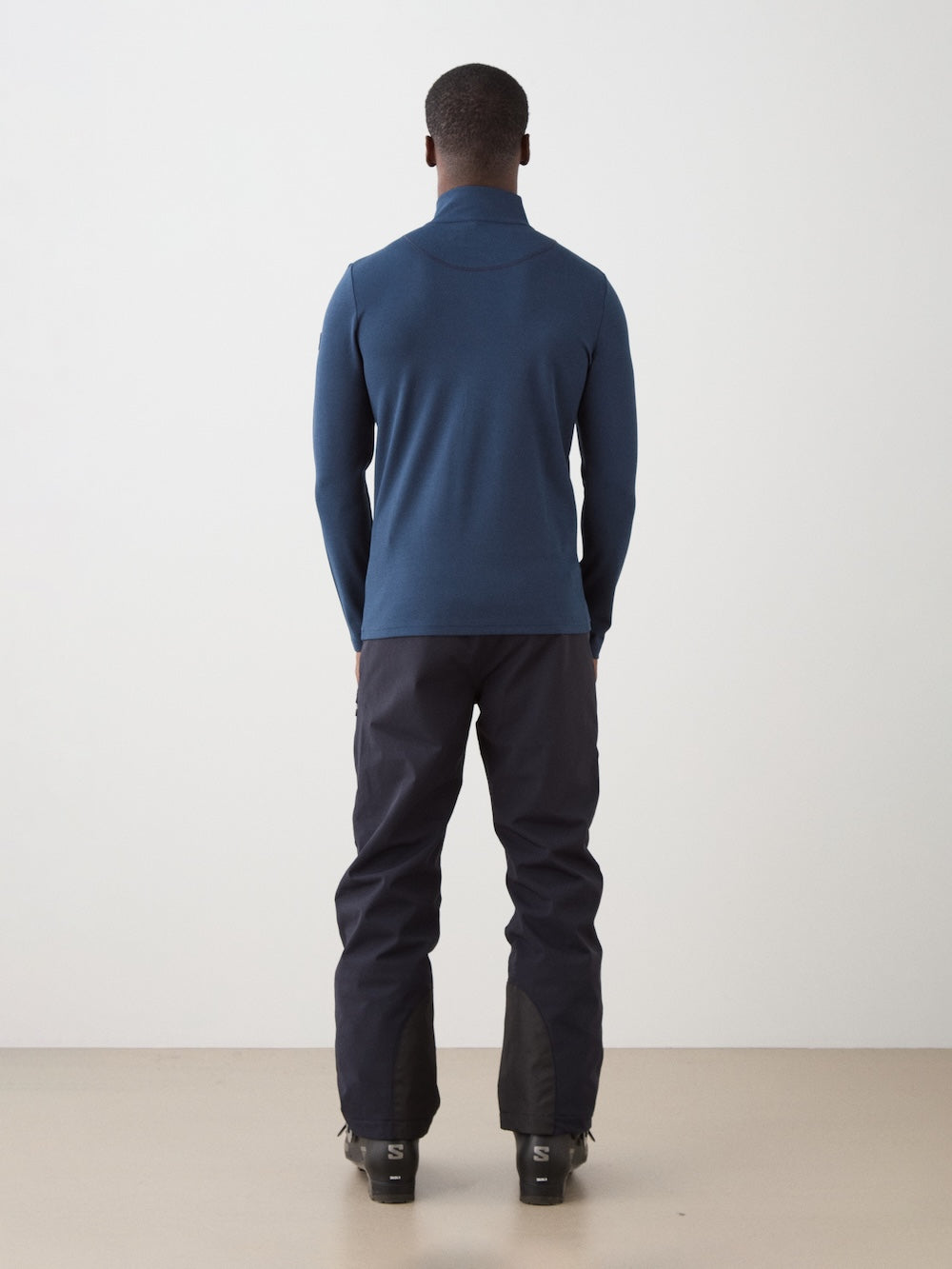 A person stands with their back to the camera, wearing the We Norwegians Moore ZipUp men long-sleeve blue turtleneck, dark pants, and black boots against a plain white wall and beige floor.