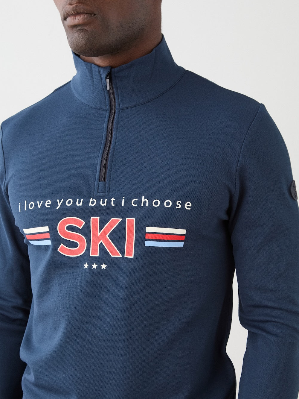 A man wearing the We Norwegians Moore ZipUp, featuring blue fabric, retro red, white, and blue ski base layer stripes on the front, and the text "i love you but i choose SKI," stands turned slightly to the side.
