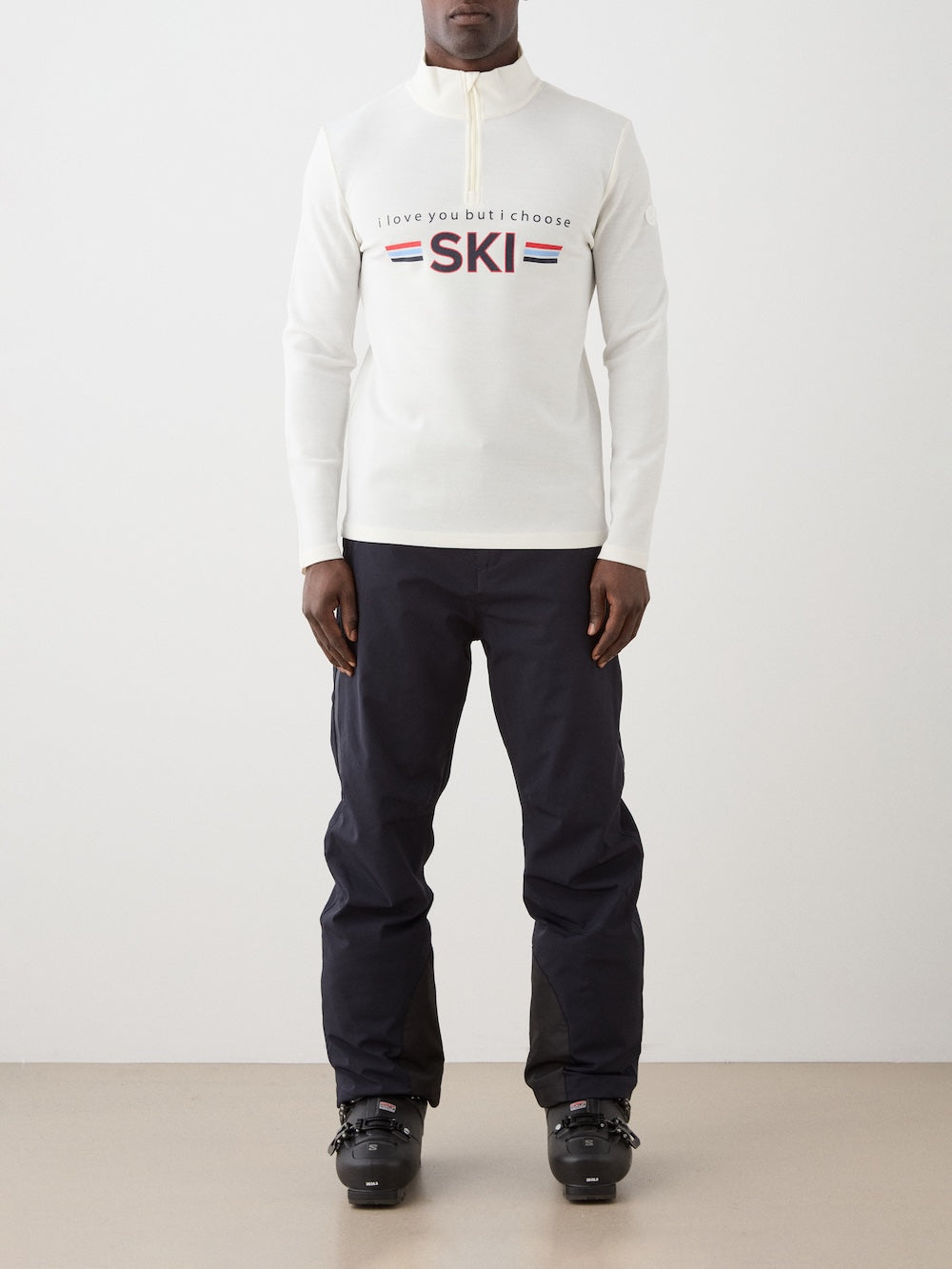 A person stands indoors wearing the We Norwegians Moore ZipUp men, a white retro ski base layer with "I love you but I choose SKI" on the front, styled with navy ski pants and black boots. The minimal background highlights this classic top.