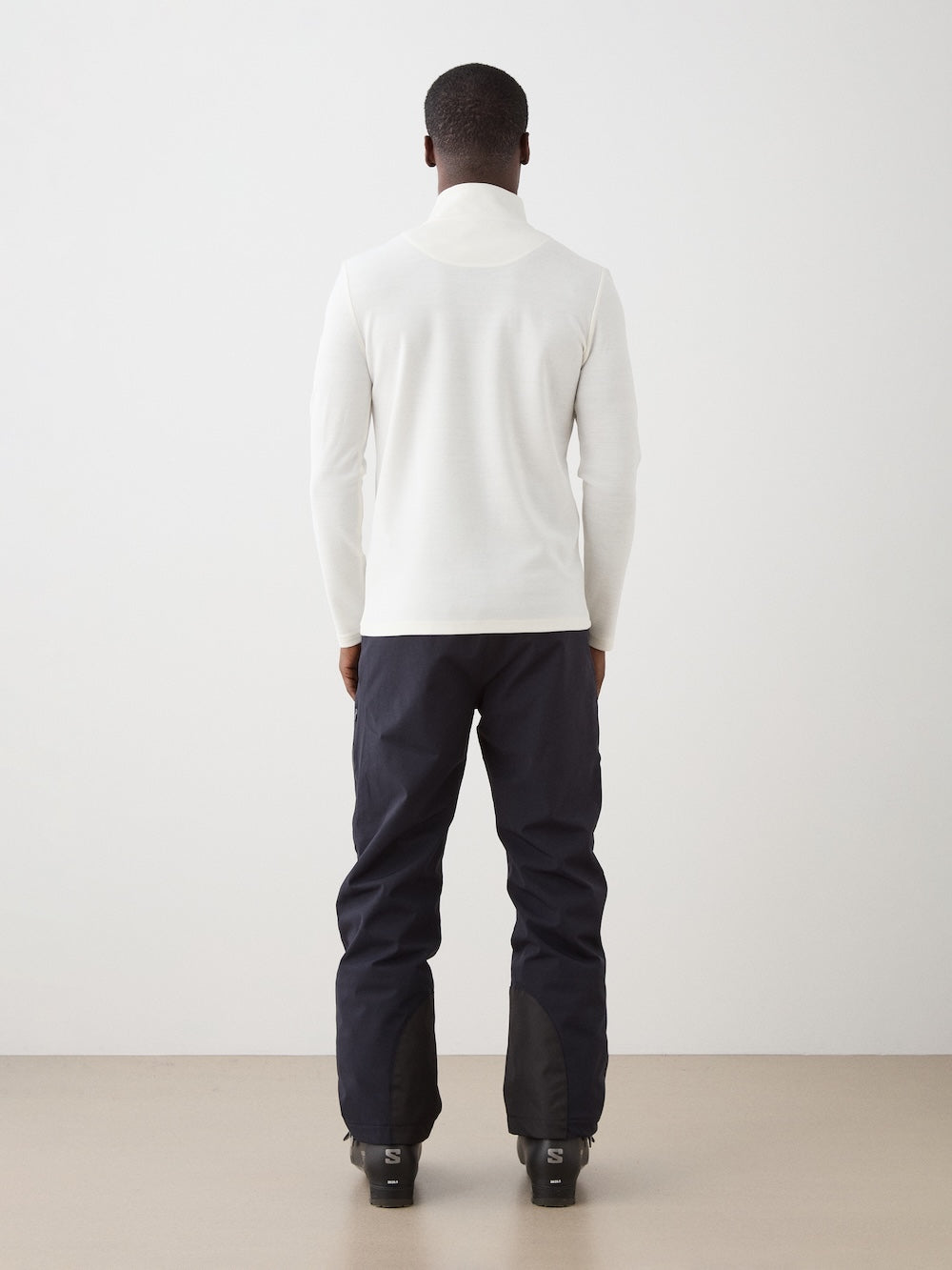 A man faces away from the camera, dressed in a white We Norwegians Moore ZipUp men’s retro ski base layer and black ski pants with gaiters over black ski boots, set against a plain light background.