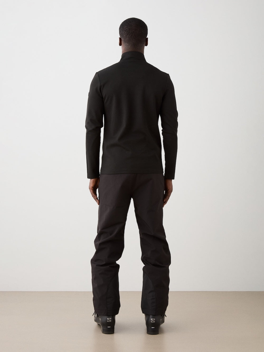 A person faces away from the camera against a plain background, wearing black alpine attire featuring the Moore ZipUp Solid men base layer by We Norwegians, paired with black pants and matching shoes.