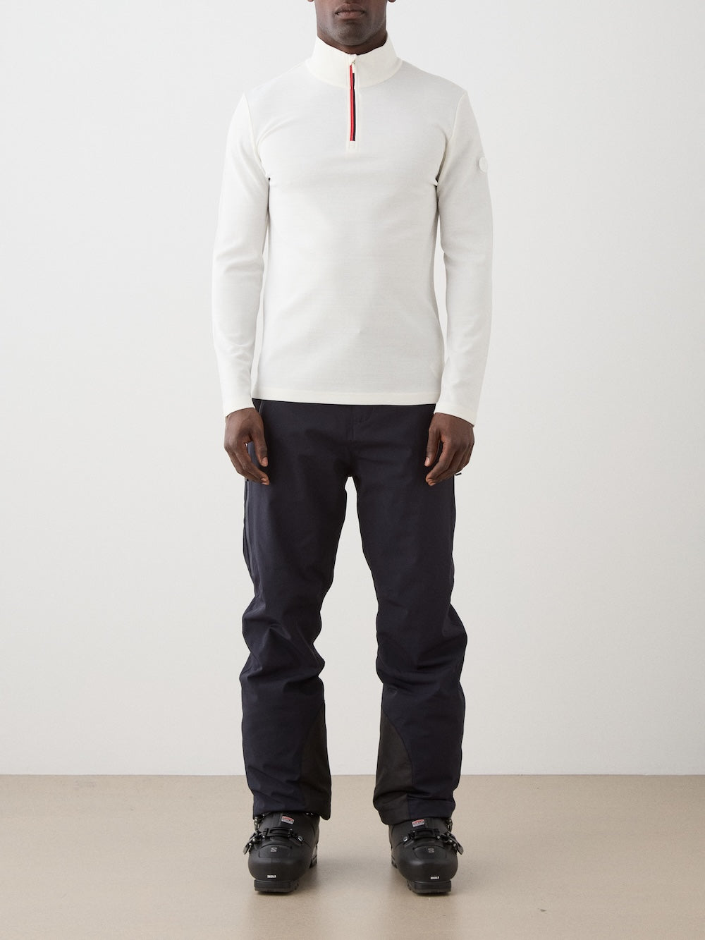 A person stands against a plain background wearing the We Norwegians Moore ZipUp Solid men’s white alpine-style long-sleeve base layer with a red zipper, paired with dark pants and black ski boots.