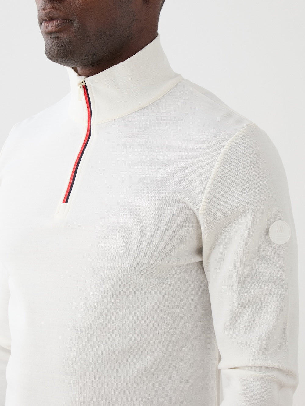 A man wears the We Norwegians Moore ZipUp Solid—a Merino wool off-white quarter-zip with a red and navy zipper stripe and a round logo patch on the sleeve—against a light, alpine-inspired background.