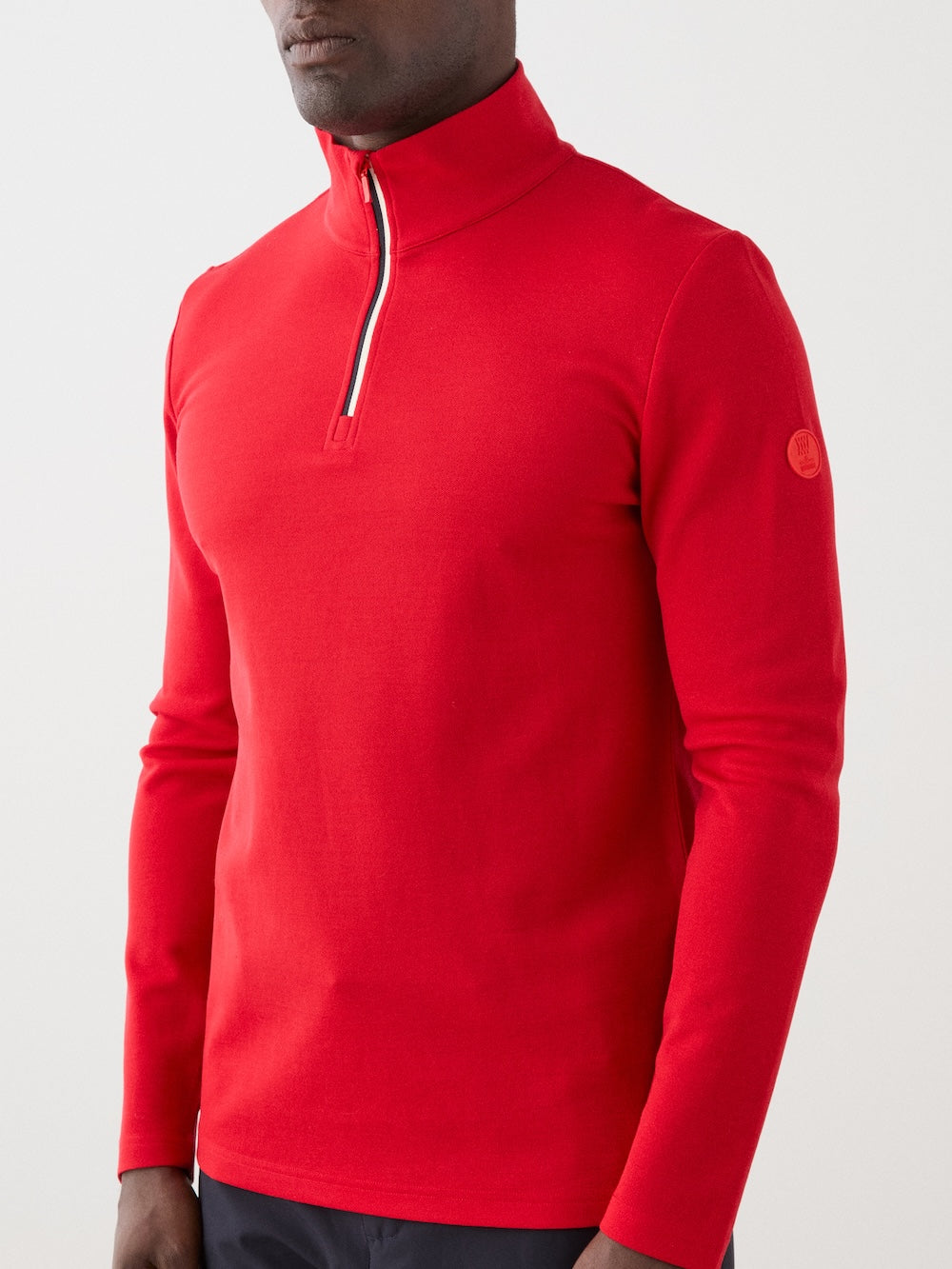 A man in a bright red We Norwegians Moore ZipUp Solid Merino wool quarter-zip pullover with long sleeves, a stand collar, and a small logo patch on the sleeve stands against a plain light background.