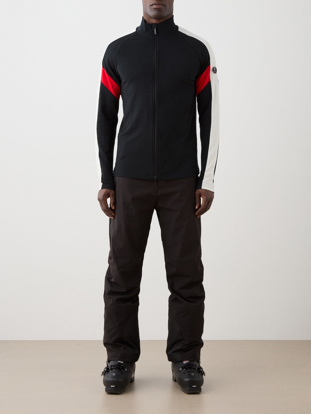 A person stands indoors against a plain background wearing the We Norwegians Zermatt FullZip men jacket, featuring a black color with a red and white stripe on the left sleeve, paired with dark pants and black ski boots.