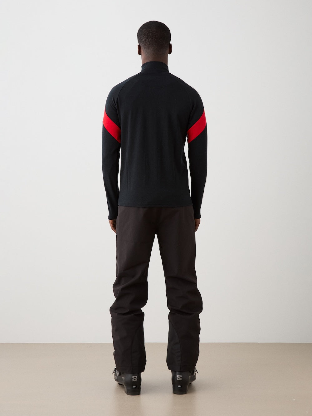 A person stands facing away from the camera, wearing the We Norwegians Zermatt FullZip men jacket with a bold red diagonal stripe on the upper sleeves, along with black pants and boots, against a plain light-colored background.