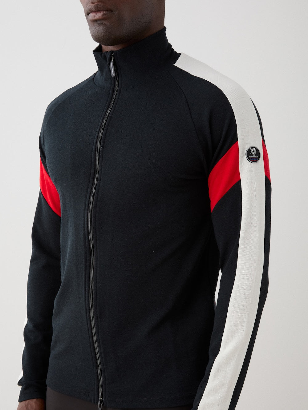 A man wearing a We Norwegians Zermatt FullZip men’s black jacket with a white stripe and red accent on the sleeves, standing against a plain light background.